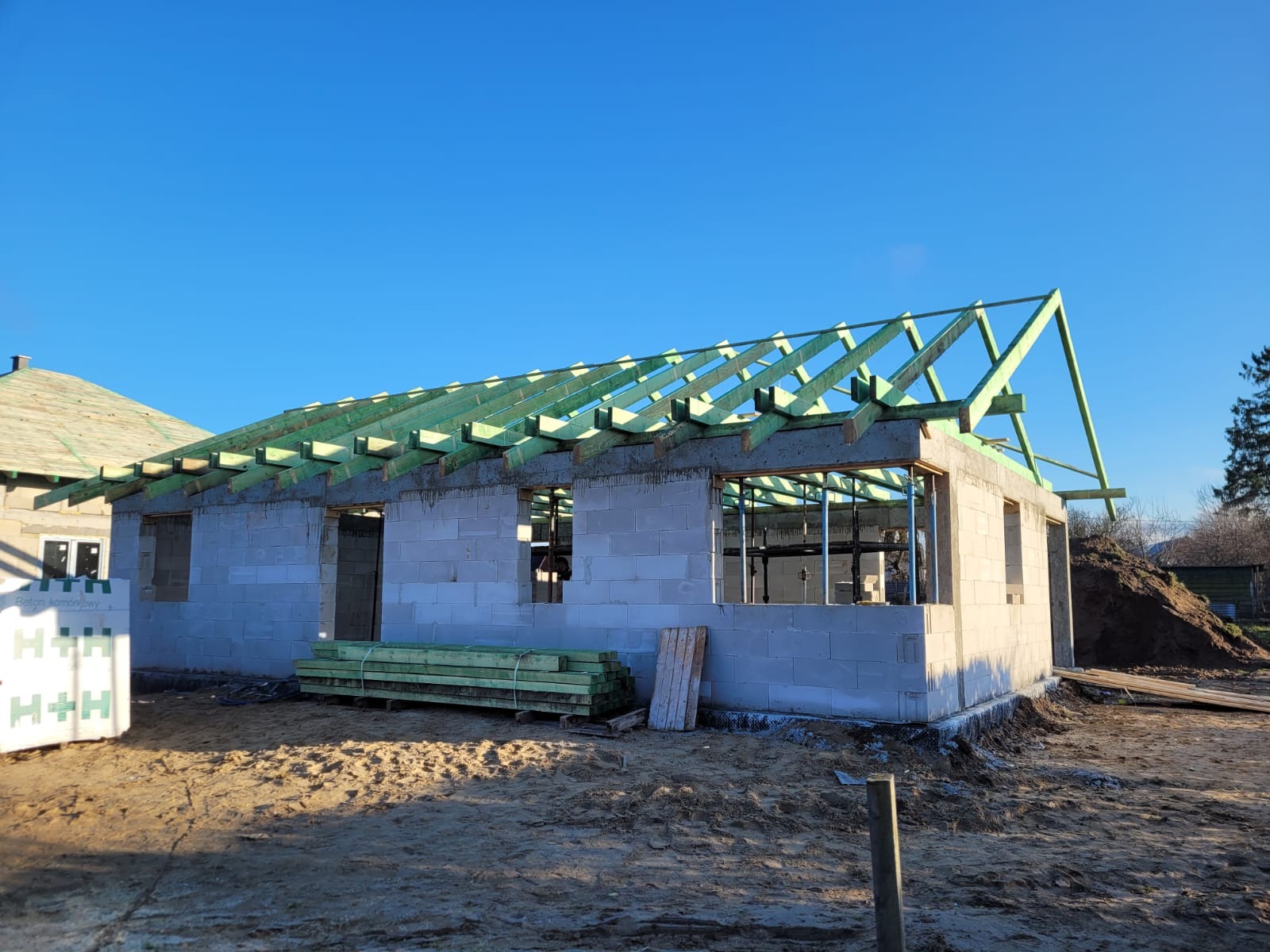 Budowa domu z betonu komórkowego w stanie surowym otwartym, widoczne ściany, więźba dachowa zaimpregnowana na zielono, paleta bloczków H+H. Słoneczny dzień.
