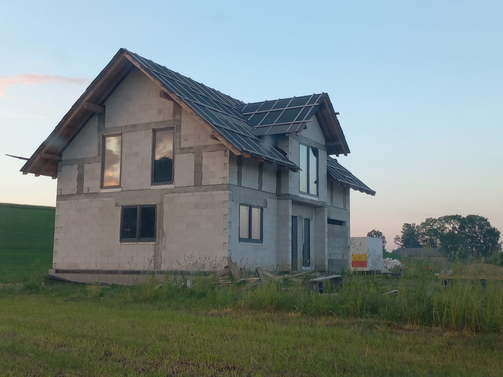 Surowy dom z betonu komórkowego w trakcie budowy, z zamontowanymi oknami i częściowo pokrytym dachem, na tle zielonej łąki i błękitnego nieba o zmierzchu.