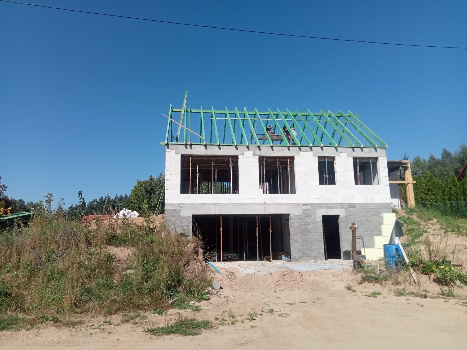 Dwukondygnacyjny dom w trakcie budowy: białe ściany z betonu komórkowego, zielona konstrukcja dachu, widoczne zbrojenia okien. Prace budowlane w toku, słoneczny dzień.