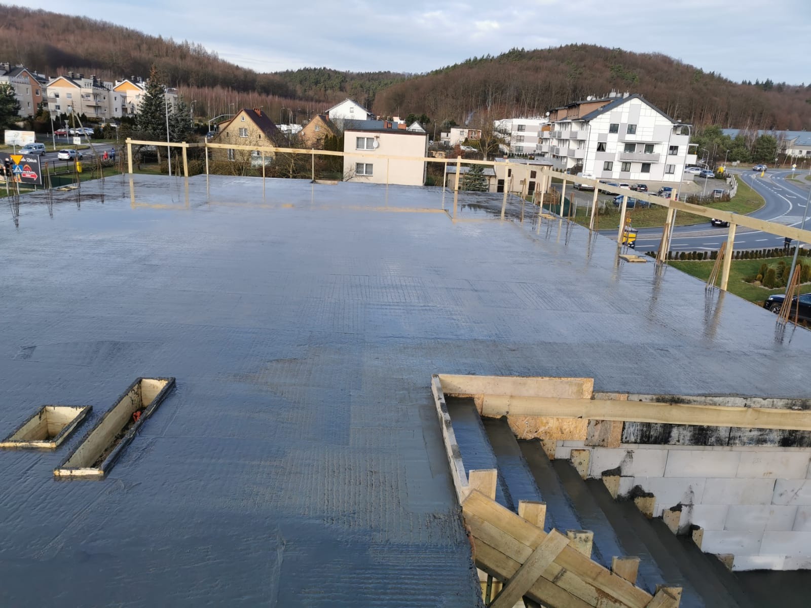 Wylany beton na stropie budynku w trakcie budowy. Widoczne szalunki i otwory w stropie. W tle zabudowa miejska i zalesione wzgórza.