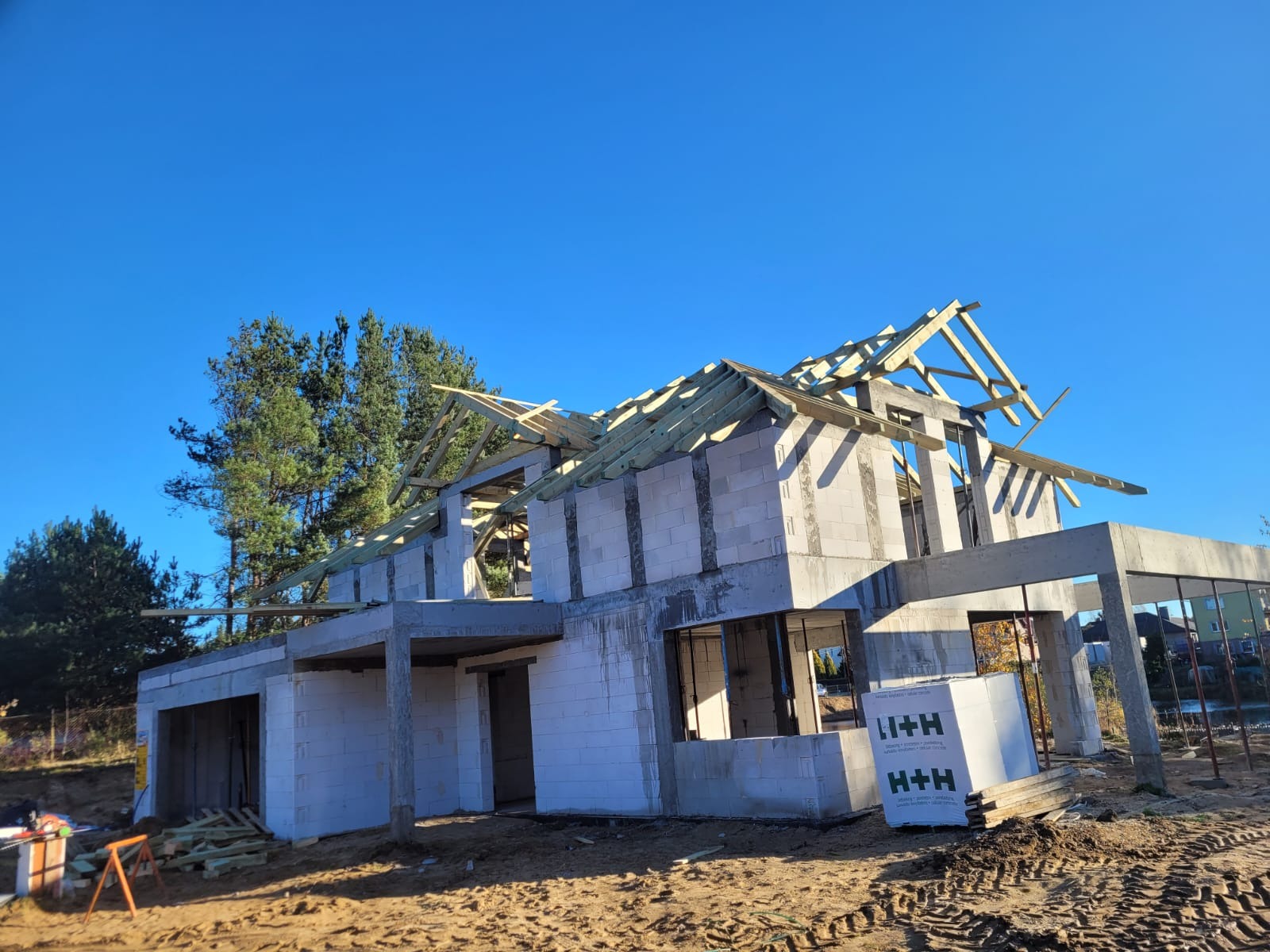 Surowy stan domu z betonu komórkowego H+H w trakcie budowy, widoczne drewniane elementy konstrukcji dachu na tle błękitnego nieba i drzew.