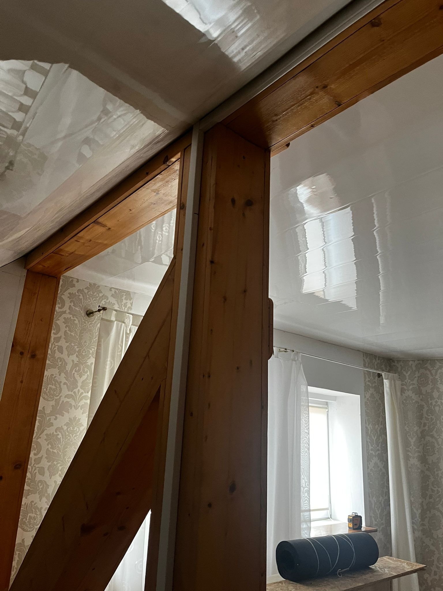 Drewniana konstrukcja stropu z widoczną błyszczącą, białą boazerią sufitową i fragmentem pokoju z oknem w tle. Ujęcie z dołu, podkreślające detale wykończenia.