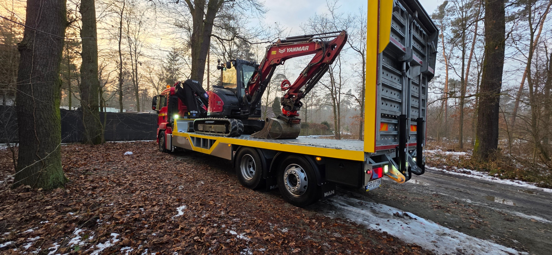 Czerwona laweta MAN z mini koparką Yanmar na platformie, zaparkowana na leśnej drodze z resztkami śniegu, w tle drzewa i zachodzące słońce.