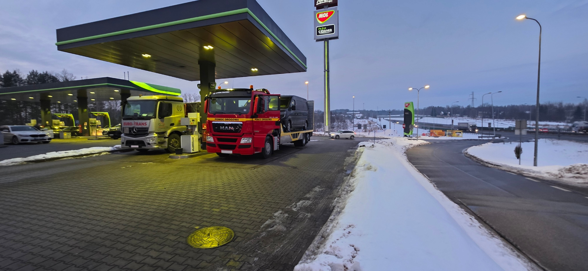 Czerwona laweta MAN z busem na platformie, stojąca na stacji benzynowej obok żółtej ciężarówki Euro-Trans, w zimowej scenerii o zmierzchu.