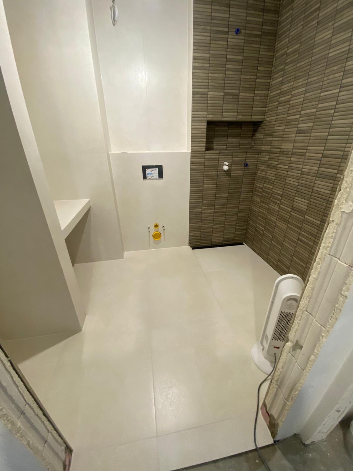 Wykończona łazienka z beżowymi płytkami imitującymi drewno pod prysznicem i jasnymi płytkami na podłodze. Widoczny grzejnik elektryczny i fragment niedokończonego wejścia.