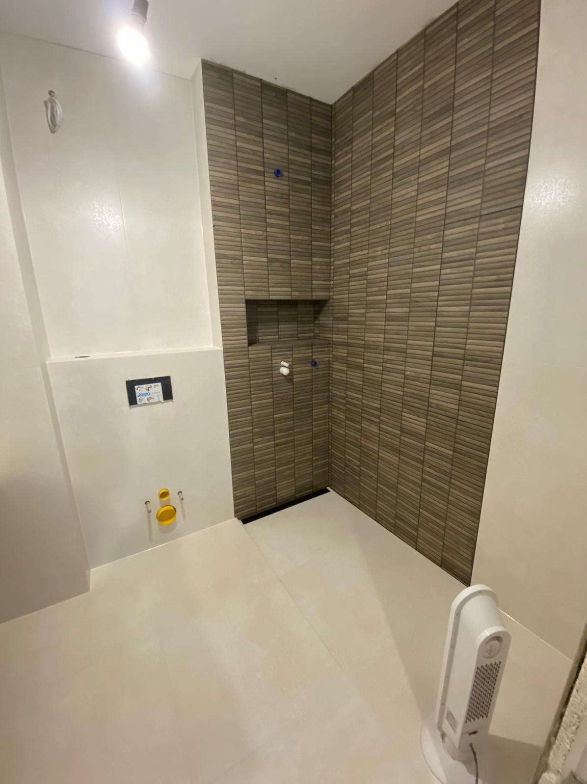 Nowoczesna łazienka po remoncie: beżowe płytki imitujące drewno w strefie prysznica, białe ściany i podłoga. Widoczne elementy instalacji i grzejnik.