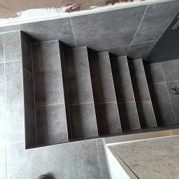 Wojciech Komar - Wykończone schody z szarych płytek, widok z góry. Widoczne buty na dole kadru. Estetyczne wykończenie krawędzi schodów listwami.