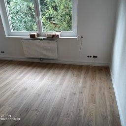 Wojciech Komar - Jasne panele podłogowe ułożone w pokoju z białymi ścianami i oknem z widokiem na zieleń. Grzejnik pod oknem, widoczne gniazdka elektryczne.