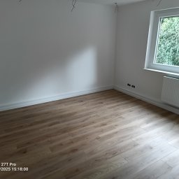 Wojciech Komar - Jasne panele podłogowe w nowym pokoju z białymi ścianami, grzejnikiem i oknem z widokiem na zieleń. Widoczne wystające kable elektryczne.