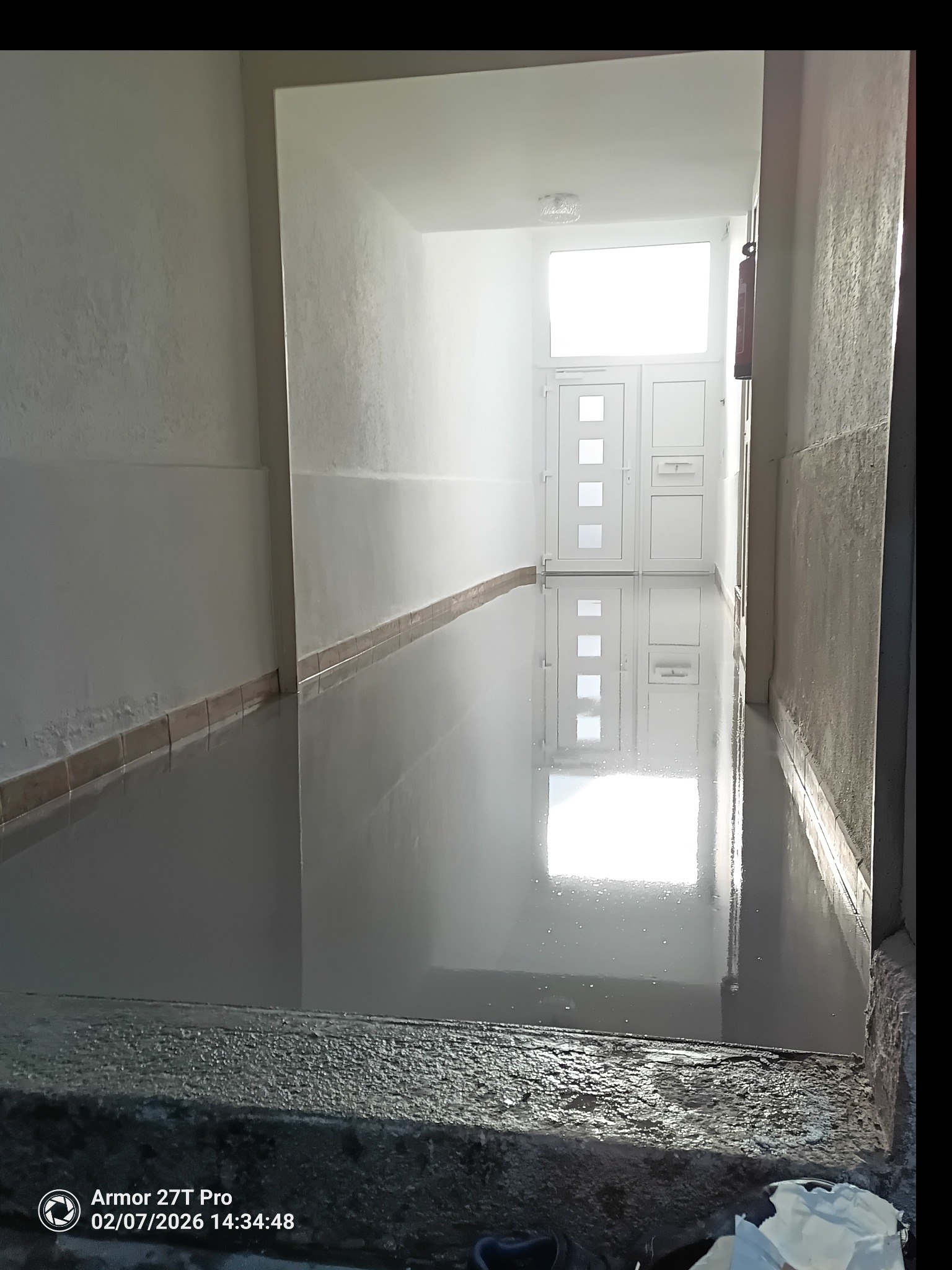 Wnętrze korytarza z wylewką samopoziomującą, odbijającą światło z okna i drzwi. Widoczne płytki przypodłogowe i ściany przygotowane do malowania.