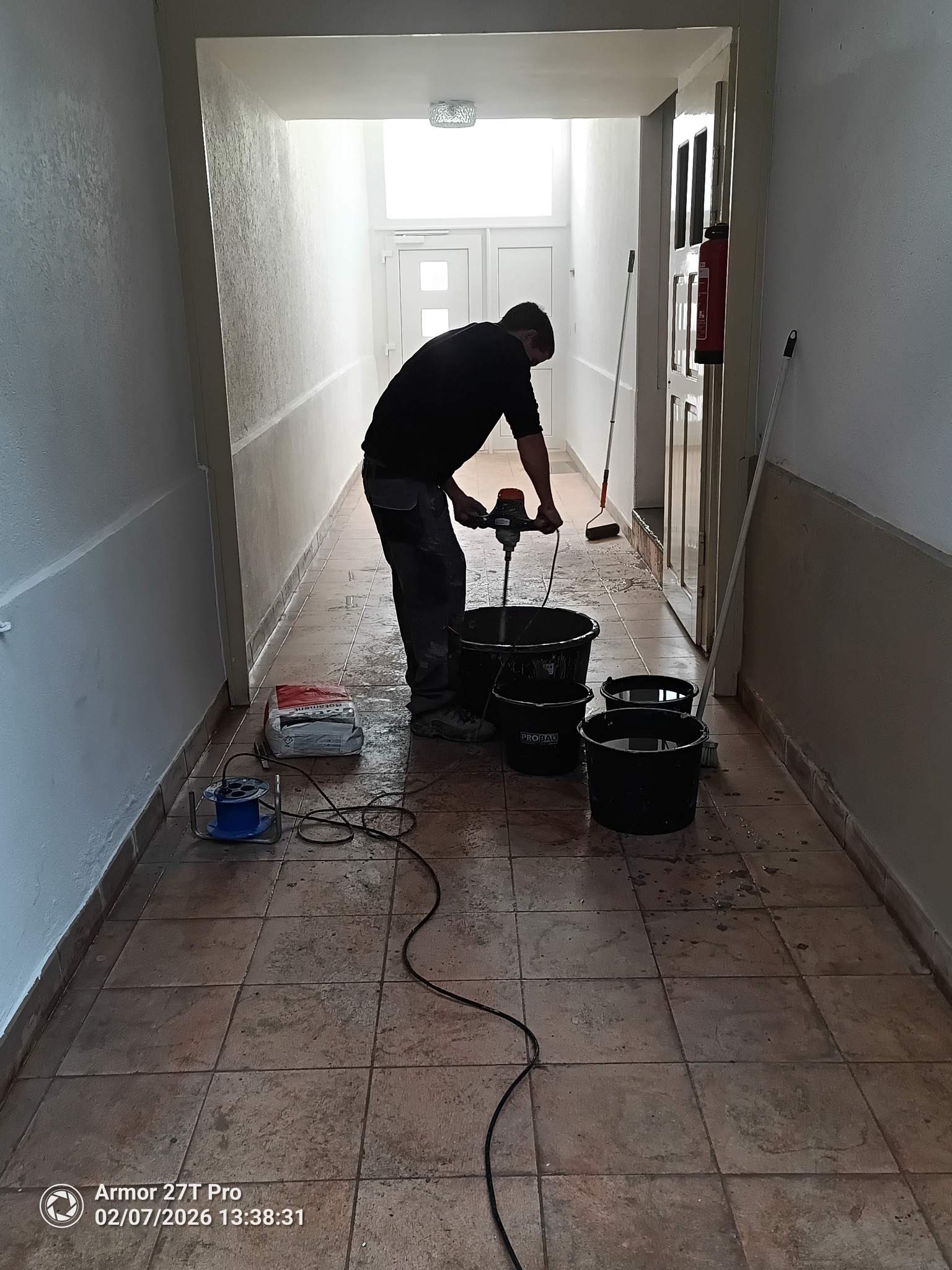 Mężczyzna miesza zaprawę w wiadrze na korytarzu z brązowymi płytkami. Widoczne wiaderka, worek z zaprawą i mikser. Przy ścianie stoi gaśnica.