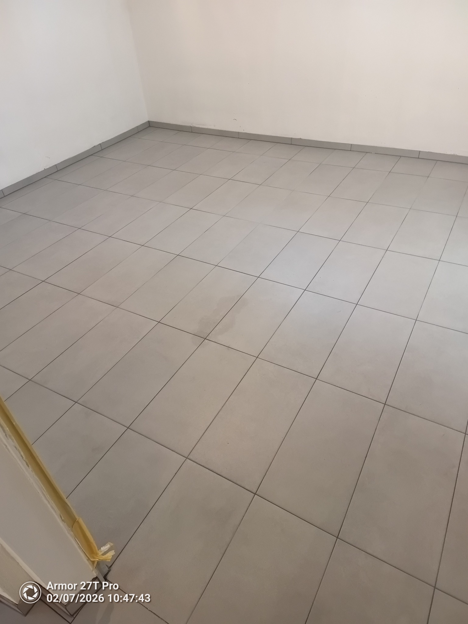 Jasnoszara, kwadratowa podłoga z płytek w pustym pomieszczeniu. Widoczne białe ściany i listwy przypodłogowe. U dołu kadru fragment drzwi z taśmą malarską.