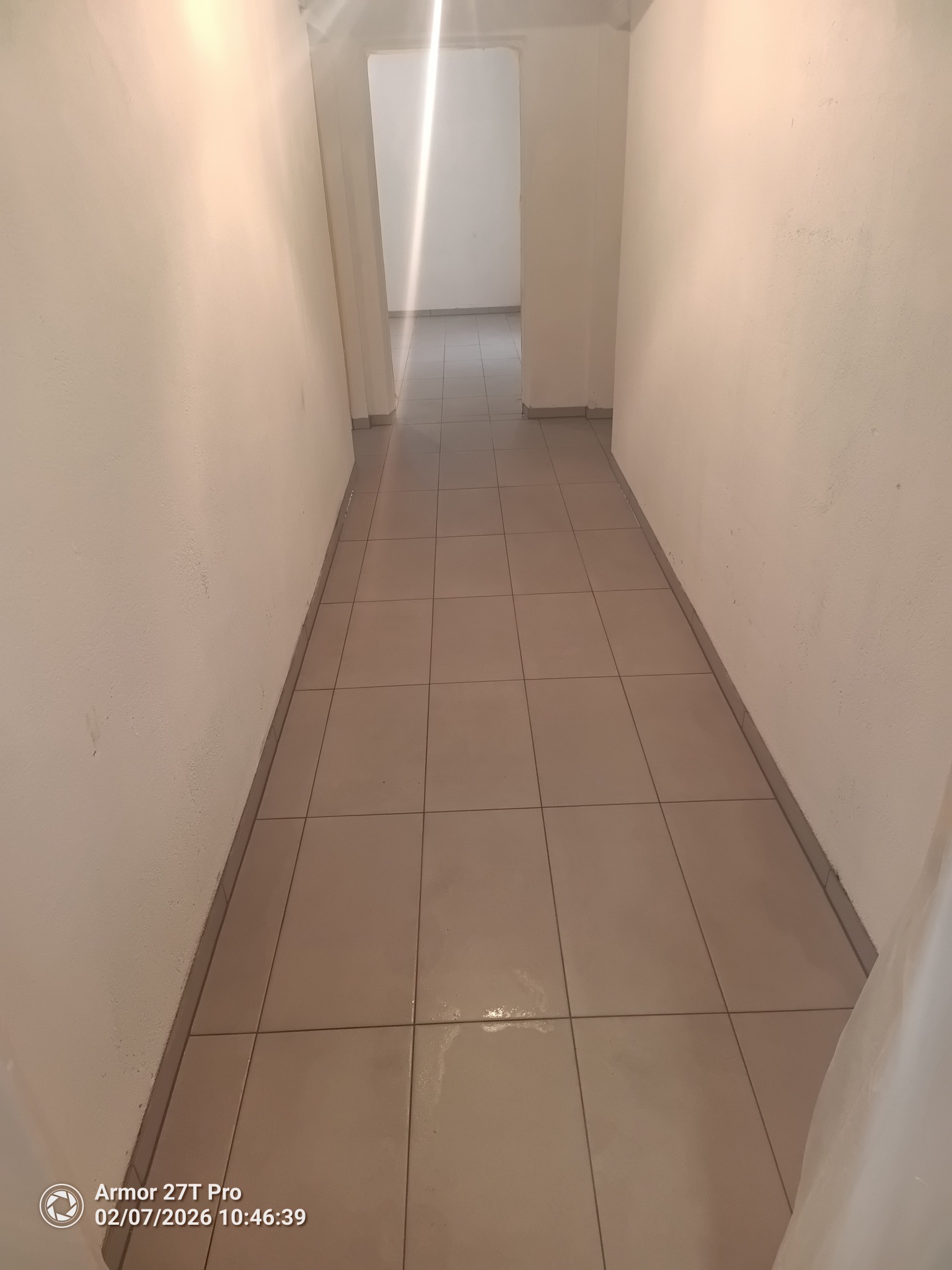 Wykończony korytarz z beżowymi płytkami podłogowymi i jasnymi ścianami. Widok w głąb, z widocznym przejściem do kolejnego pomieszczenia. Minimalistyczny styl.