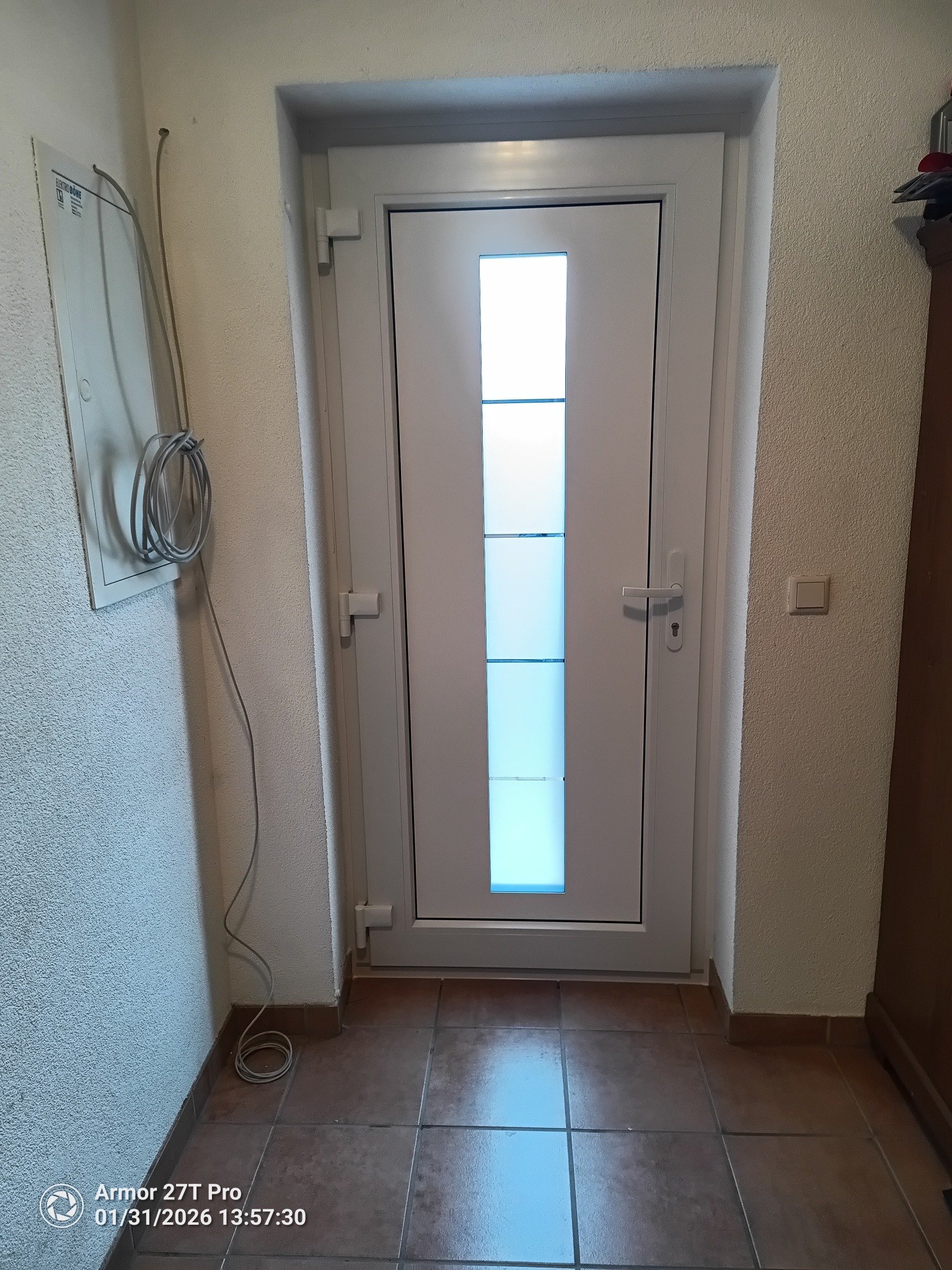 Białe drzwi wejściowe z przeszkleniami w korytarzu, brązowe płytki podłogowe, widoczna instalacja elektryczna na ścianie. Ujęcie z niskiego kąta.