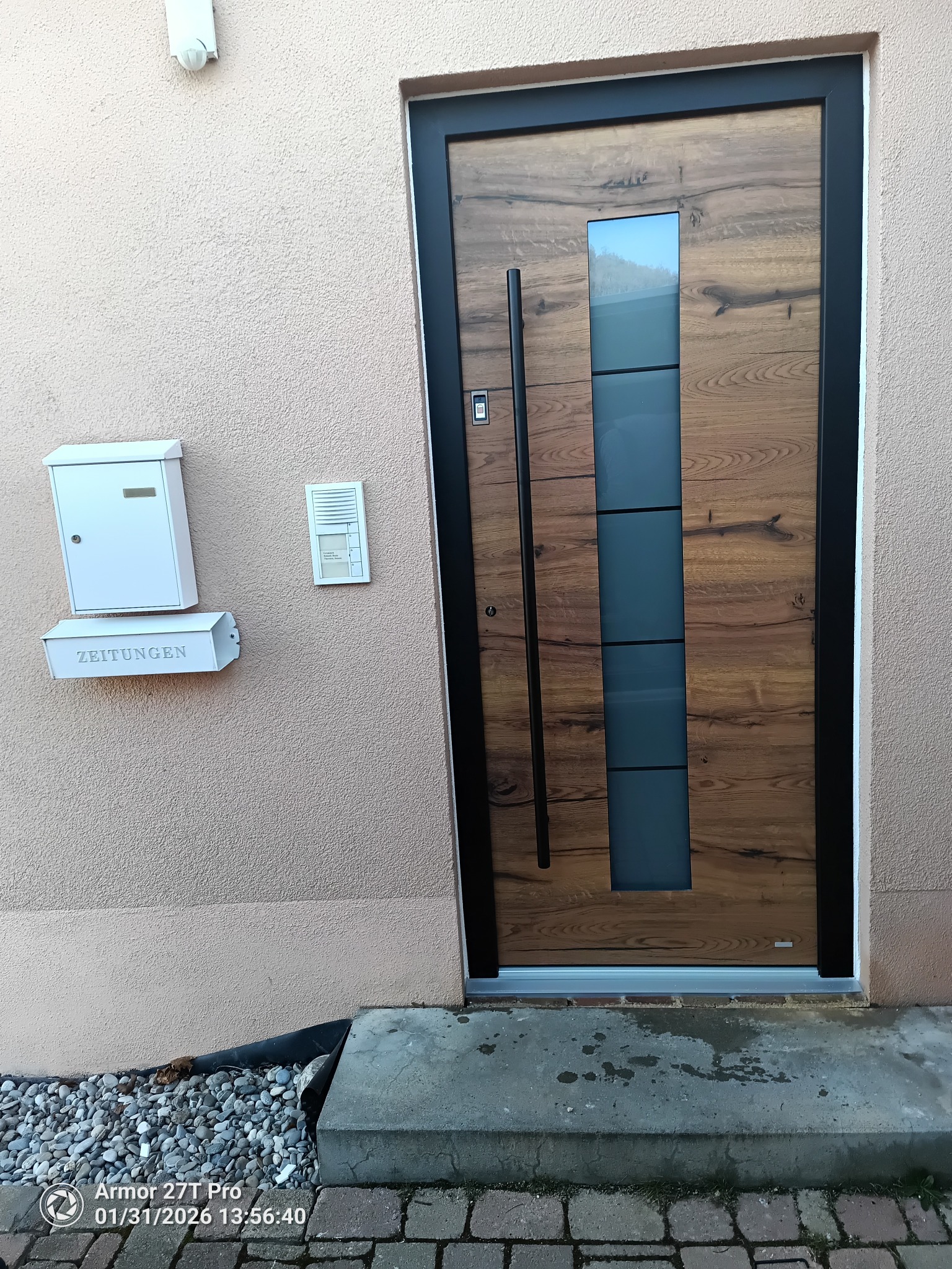 Drewniane drzwi wejściowe z czarną ramą i szybą, zamontowane w beżowej ścianie obok skrzynki na listy i domofonu. Widoczne kamienie i kostka brukowa u podstawy.
