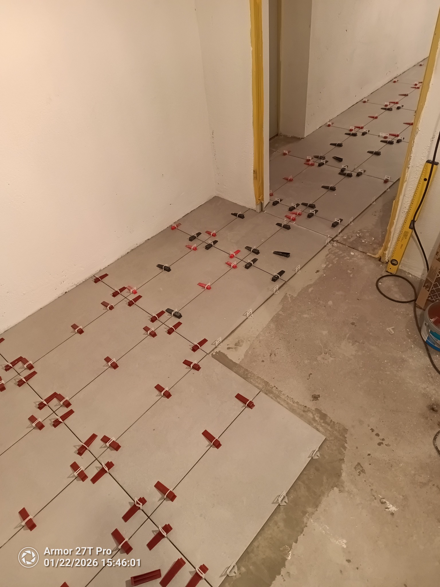 Układanie jasnych płytek podłogowych z użyciem czerwonych i czarnych klipsów poziomujących w długim korytarzu. Widoczna poziomica przy ścianie.