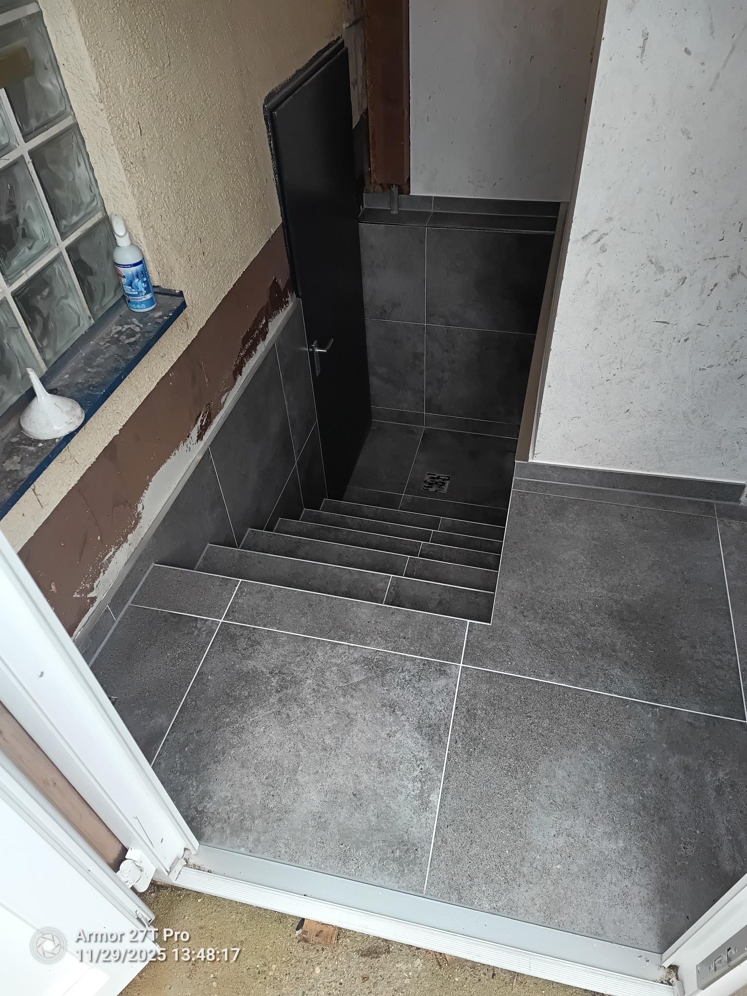 Wykończone schody w szarości, z ciemnymi płytkami i jasnymi fugami. Widok z góry, z otwartymi drzwiami i oknem z luksferami w tle. Minimalistyczny design.