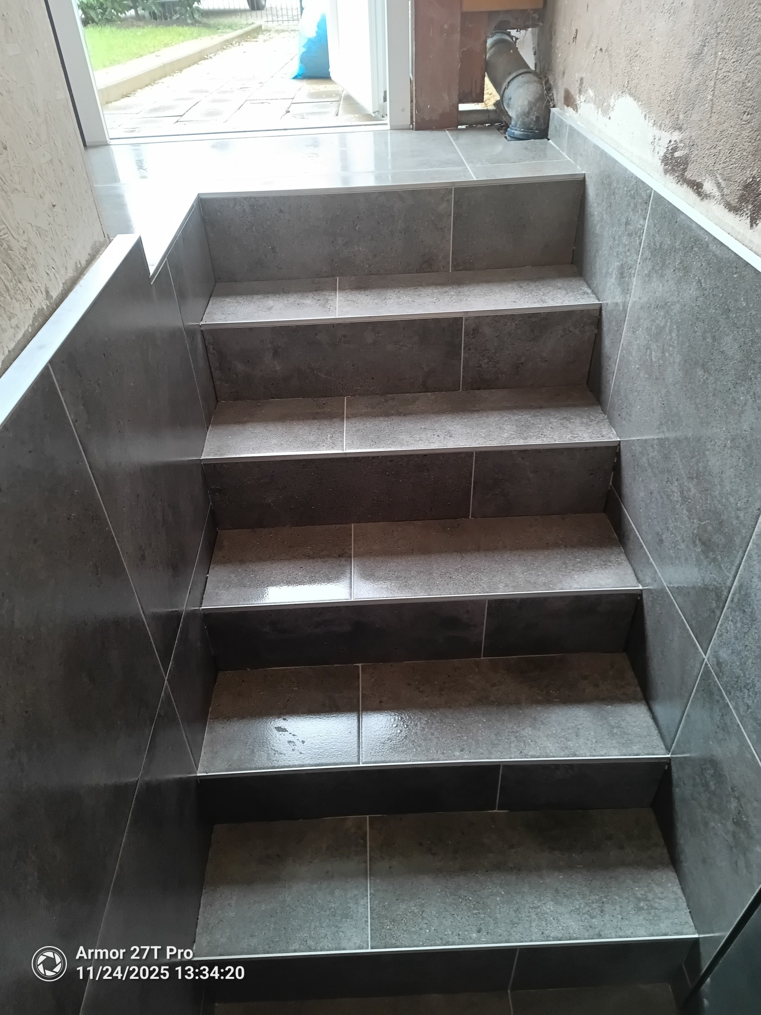 Wykończone schody z szarych płytek, widok z góry. Płytki ułożone na stopniach i ścianach. Widoczne detale wykończenia i fuga. Na górze widoczny fragment rury.