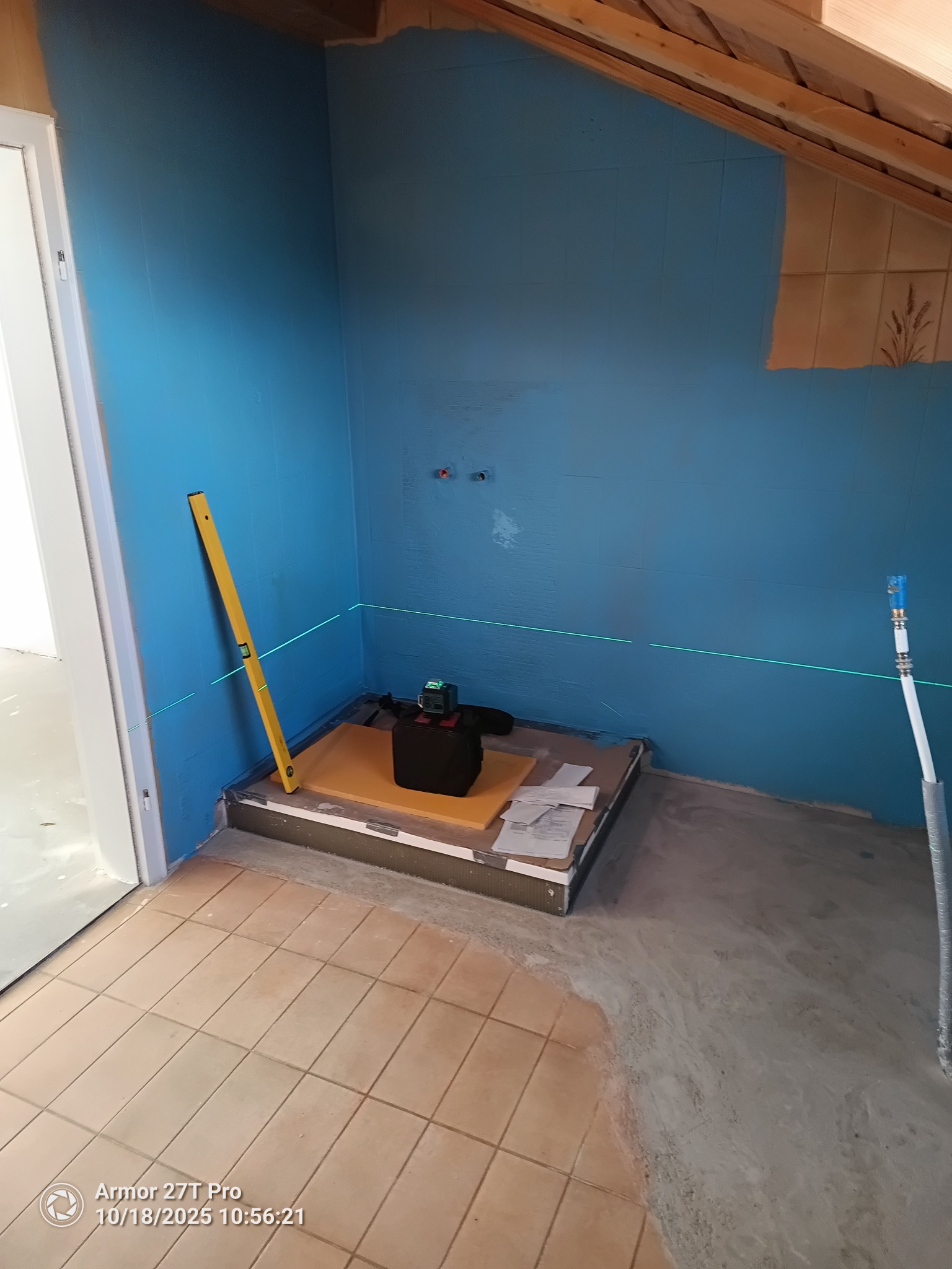 Niebieska hydroizolacja ścian w łazience, poziomica laserowa, przygotowany brodzik. Widoczne płytki na ścianie w górnym rogu. Prace przygotowawcze przed wykończeniem.