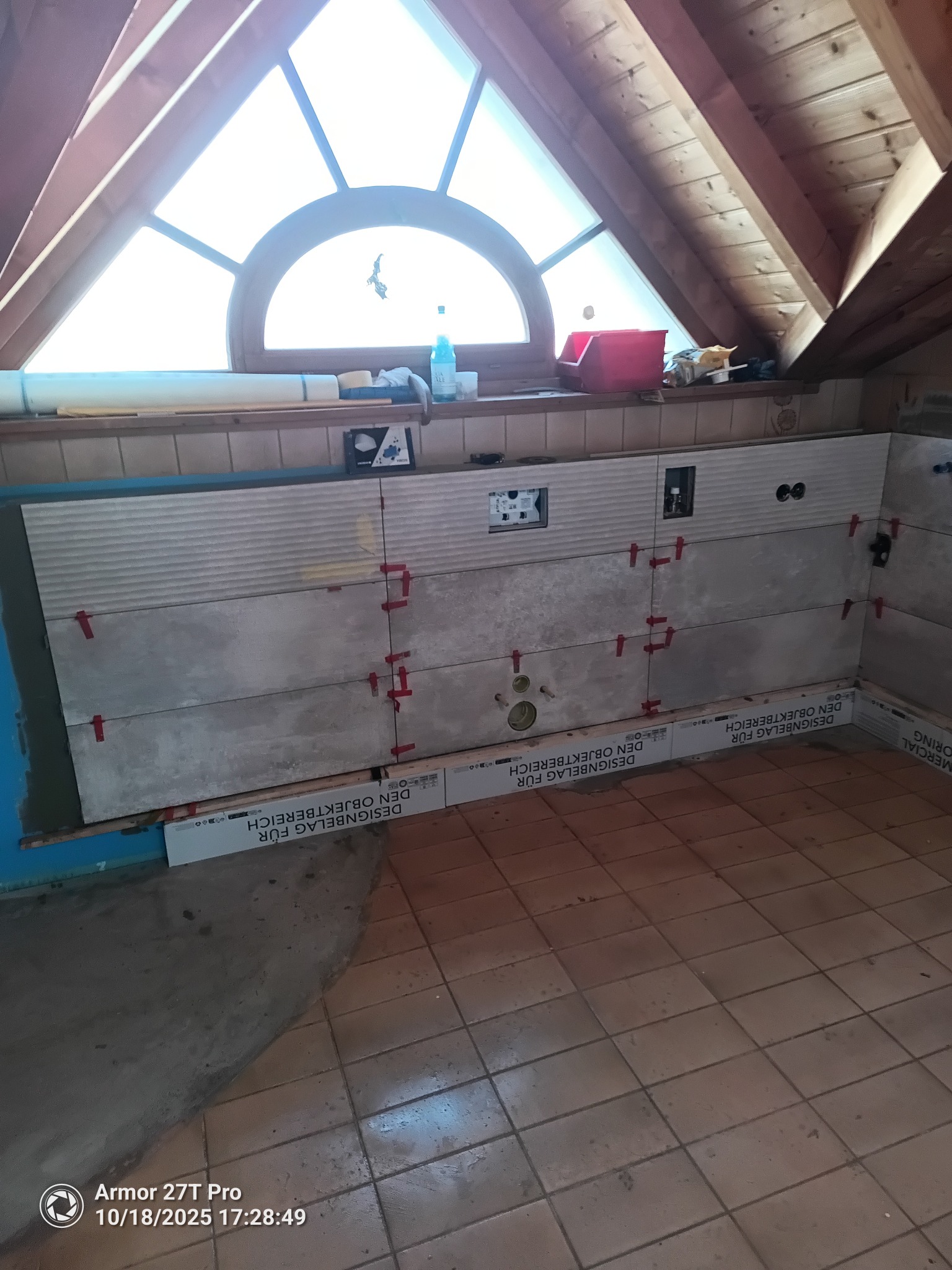 Montaż płytek ściennych w łazience na poddaszu, widoczne czerwone krzyżyki dystansowe, przygotowane otwory na instalacje. Podłoga wyłożona starą terakotą.