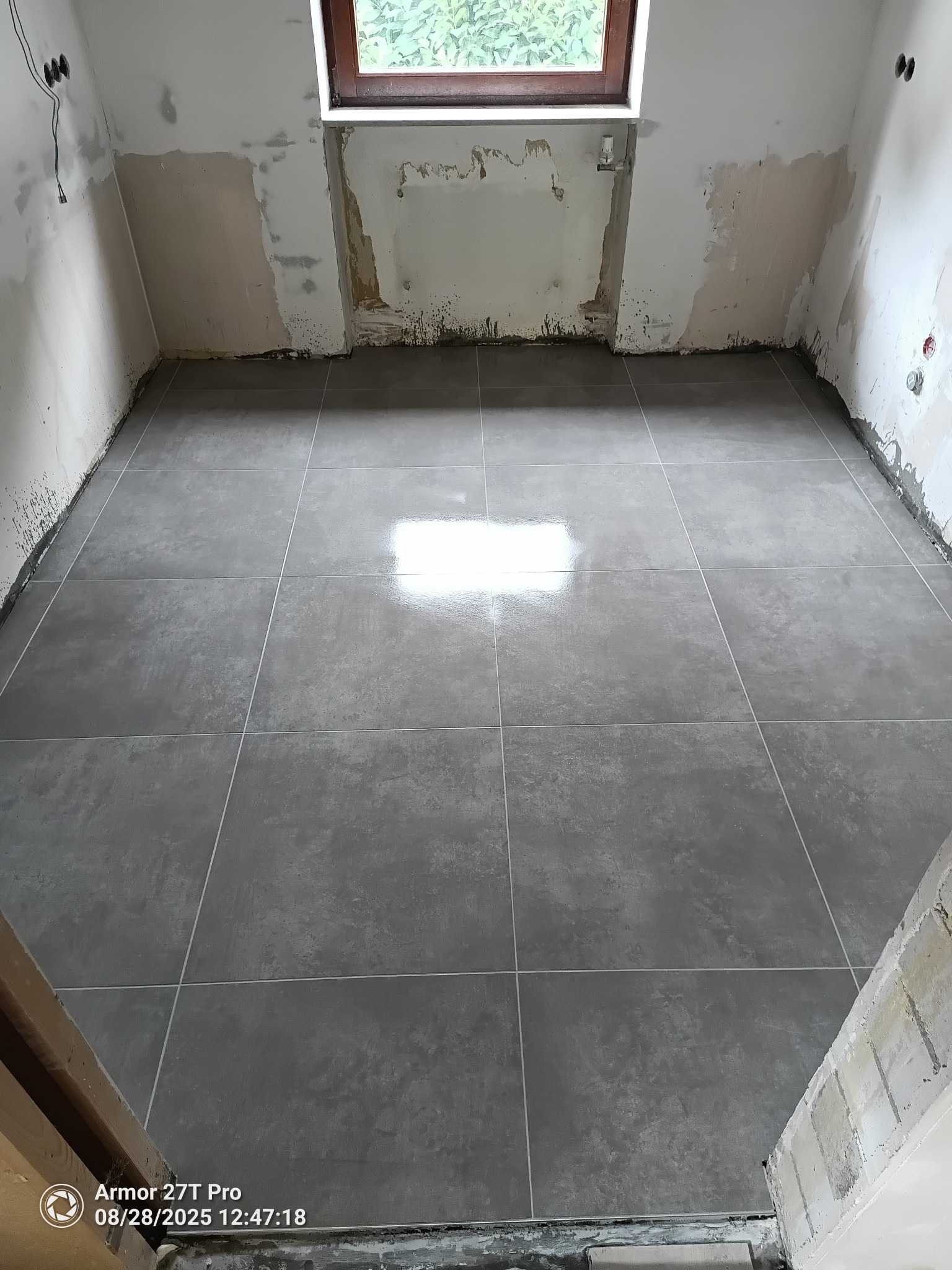 Ułożone szare płytki podłogowe w remontowanym pomieszczeniu z oknem. Ściany przygotowane do dalszych prac wykończeniowych. Widok z góry na nową podłogę.