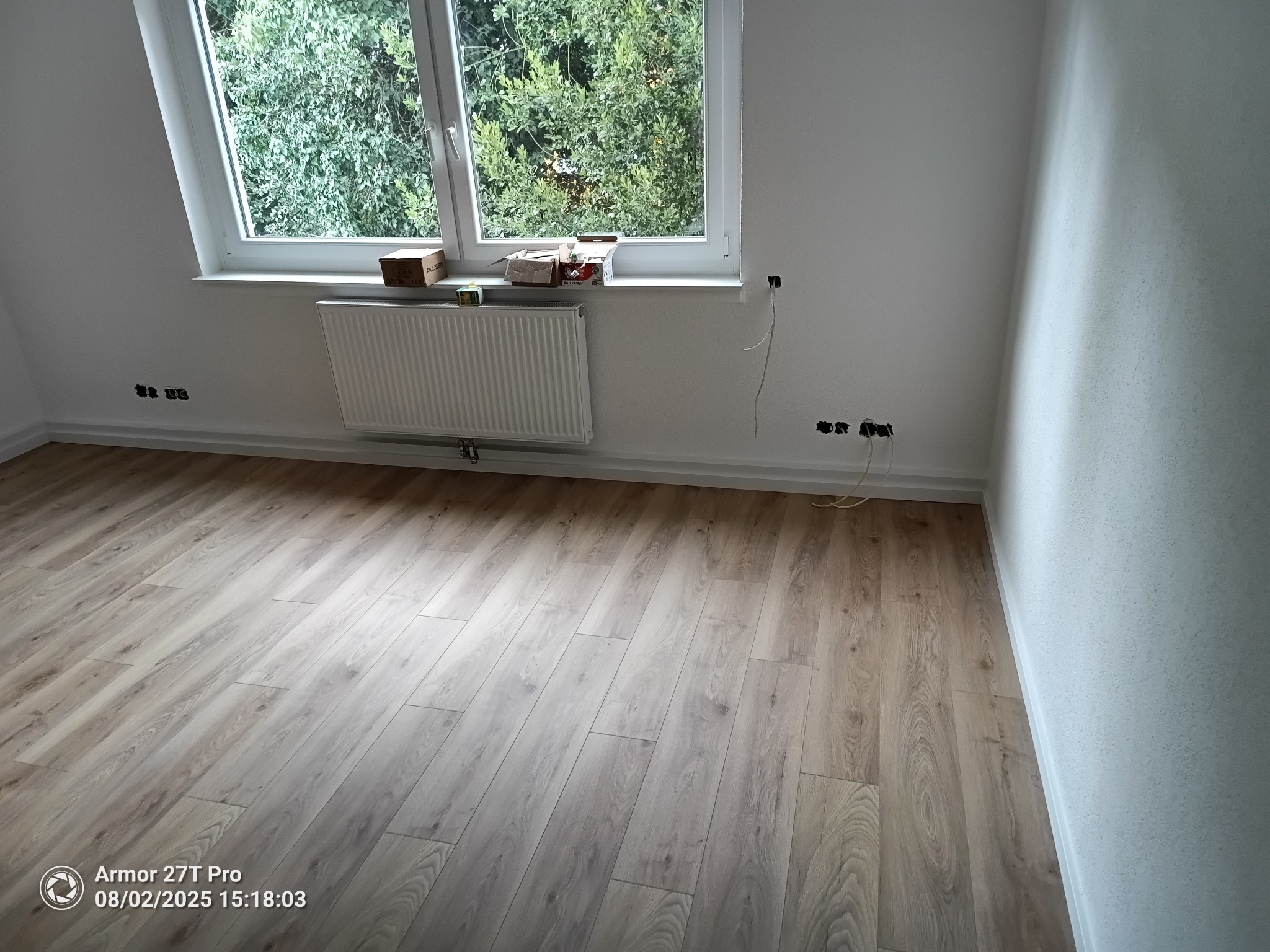 Jasne panele podłogowe ułożone w pokoju z białymi ścianami i oknem z widokiem na zieleń. Grzejnik pod oknem, widoczne gniazdka elektryczne.