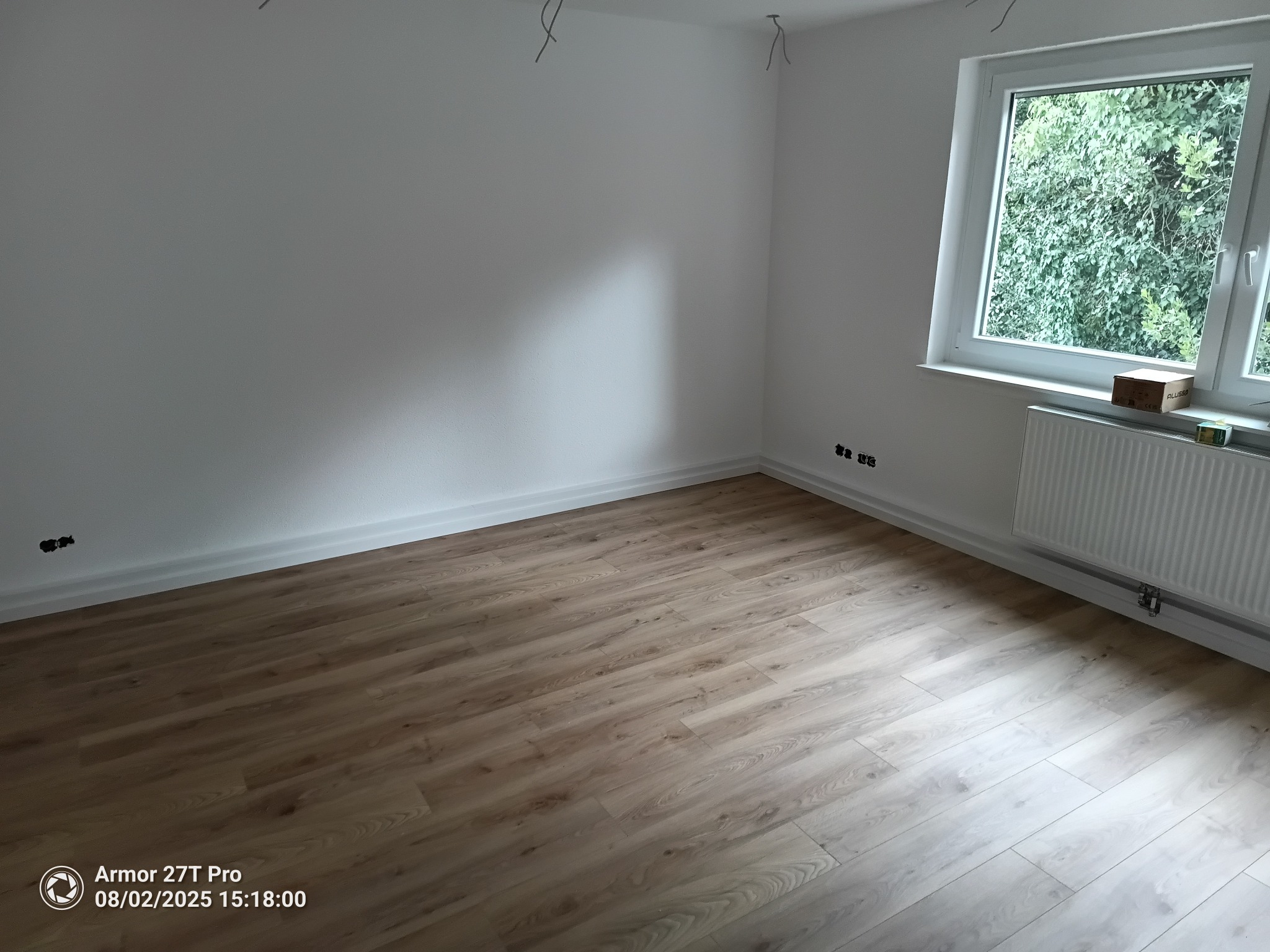 Jasne panele podłogowe w nowym pokoju z białymi ścianami, grzejnikiem i oknem z widokiem na zieleń. Widoczne wystające kable elektryczne.