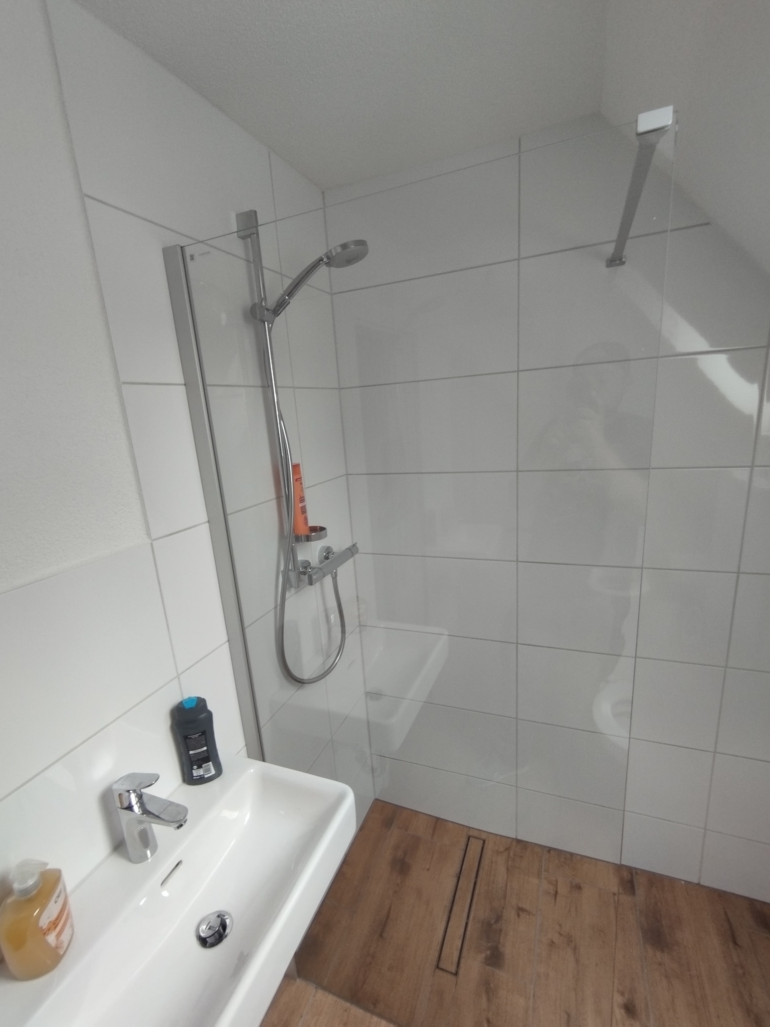 Nowoczesna kabina prysznicowa z białymi płytkami i drewnopodobną podłogą. Widoczna umywalka z chromowaną baterią i akcesoriami łazienkowymi.