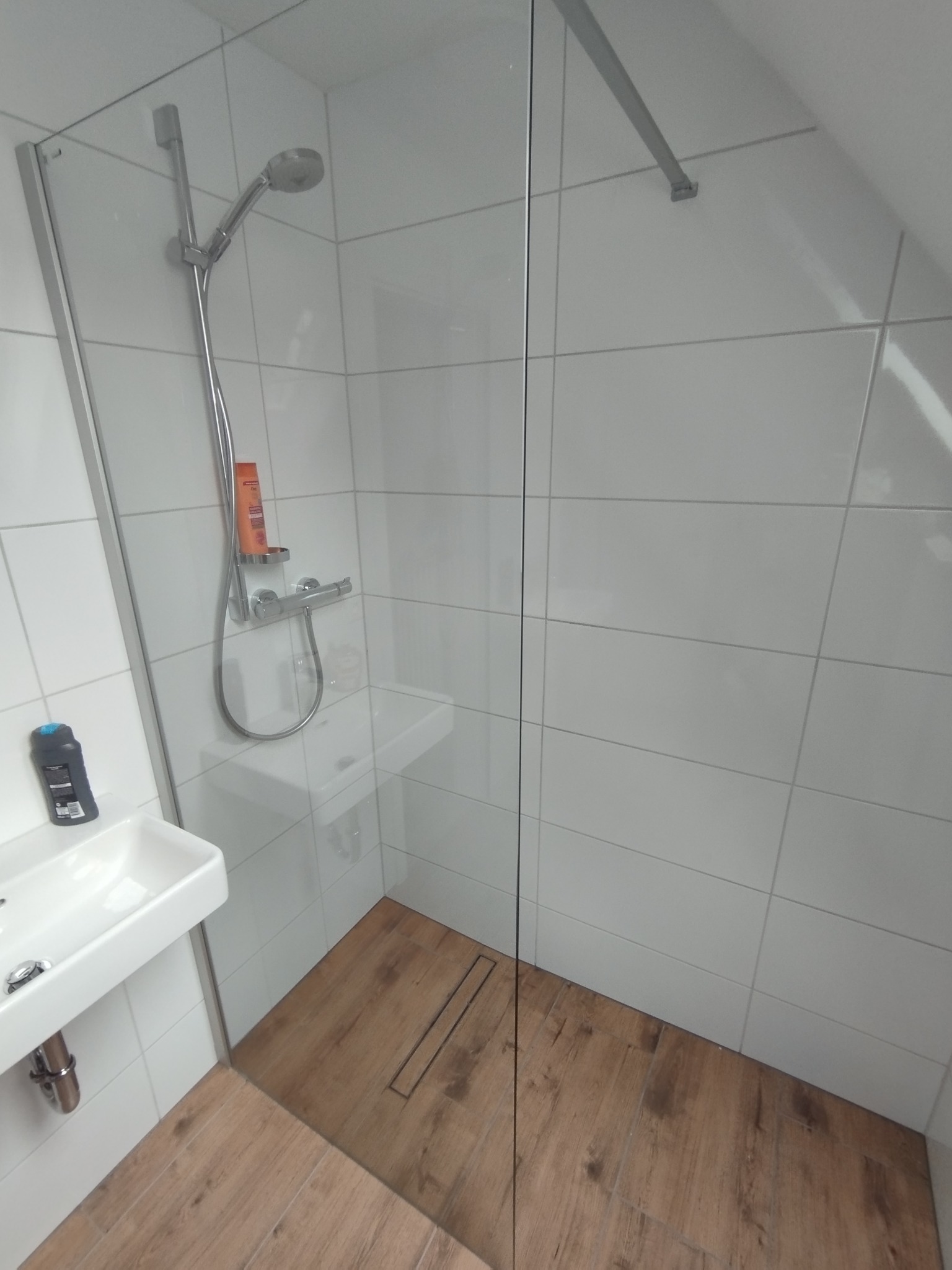 Nowoczesna kabina prysznicowa z drewnopodobną podłogą i białymi płytkami ściennymi. Widoczna szklana ściana kabiny, bateria i kosmetyki na półce. Ujęcie z perspektywy.