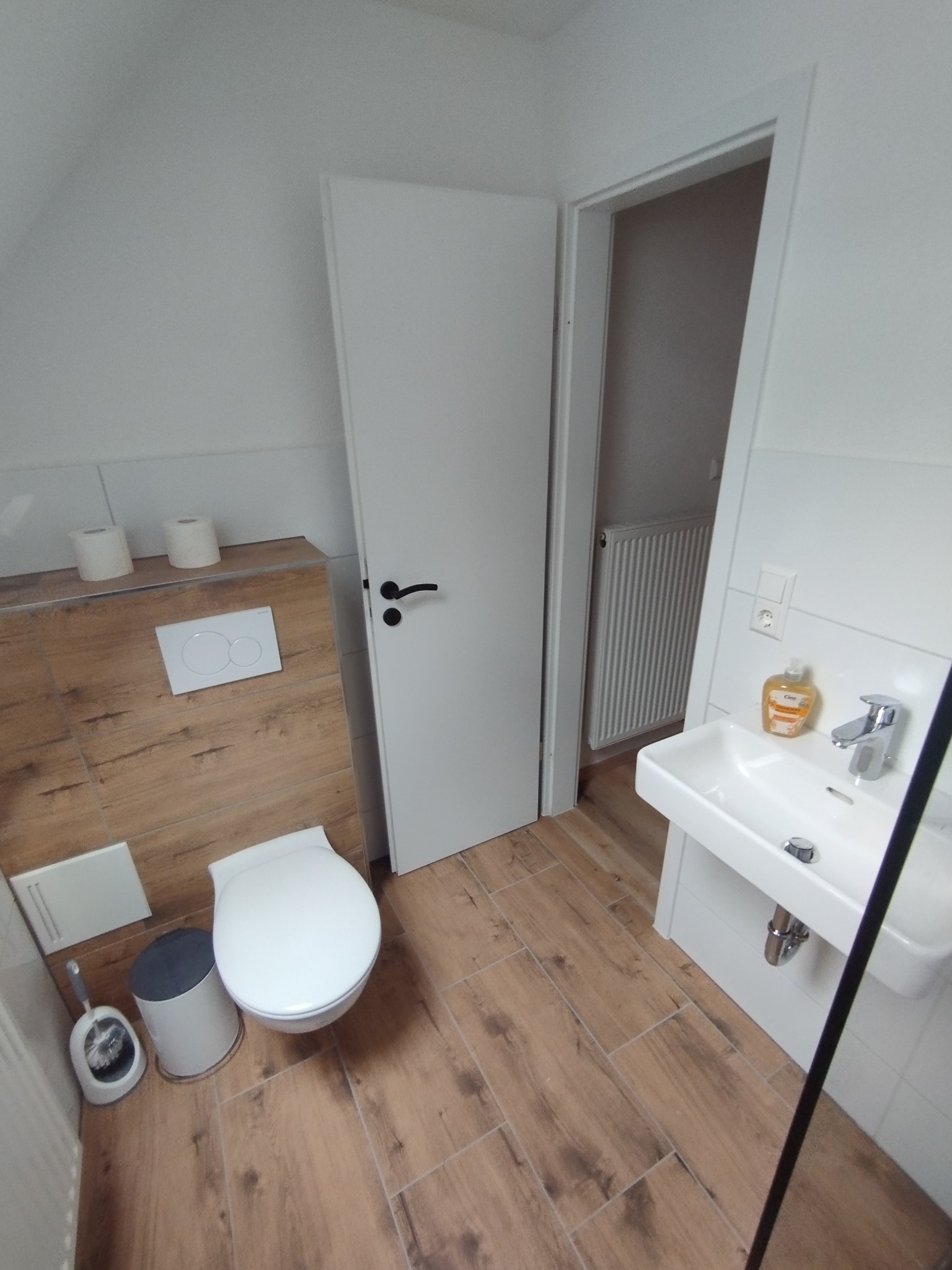 Nowoczesna łazienka na poddaszu z drewnopodobnymi płytkami, białą armaturą i drzwiami. Widoczny fragment kabiny prysznicowej.