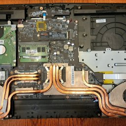 MMSerwis - Otwarty laptop z widocznymi komponentami: dysk twardy, pamięć RAM, procesor, system chłodzenia z miedzianymi rurkami cieplnymi i wentylatory. Widok z góry na drewnianym stole.