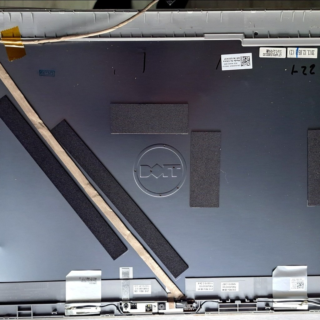 Wnętrze obudowy laptopa Dell z widocznymi elementami elektroniki, taśmami i naklejkami. Widoczne logo Dell na metalowej powierzchni.