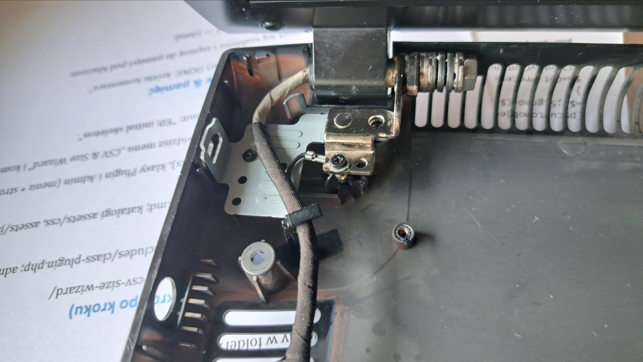 Detal naprawy laptopa: widoczne zawiasy, kabel, śruby i fragment obudowy. Elementy po demontażu, przygotowane do naprawy lub wymiany.