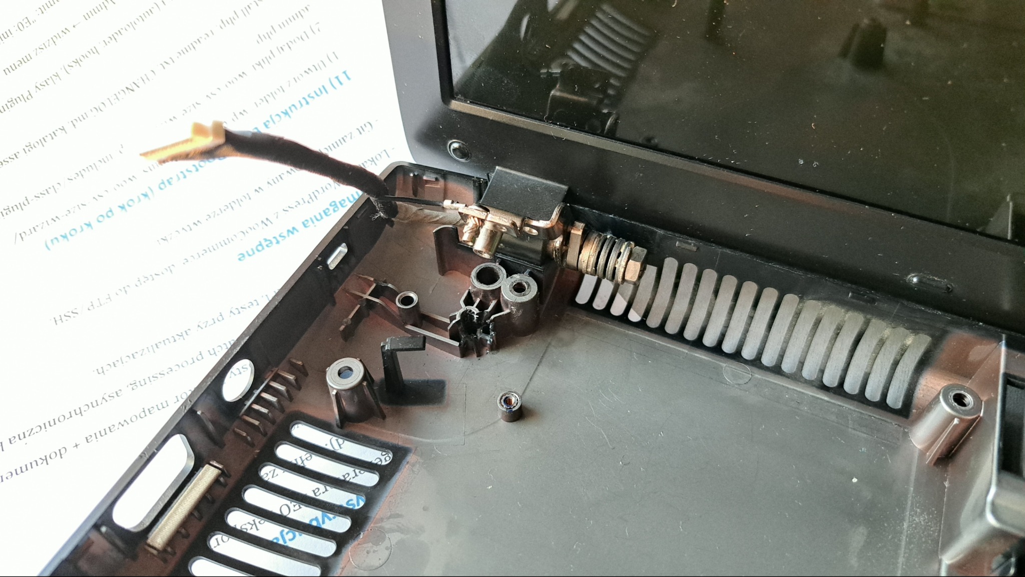 Wnętrze obudowy laptopa z widocznymi elementami mocowania zawiasu i przewodami. Widoczne ślady naprawy i demontażu. Dokument z instrukcją w tle.