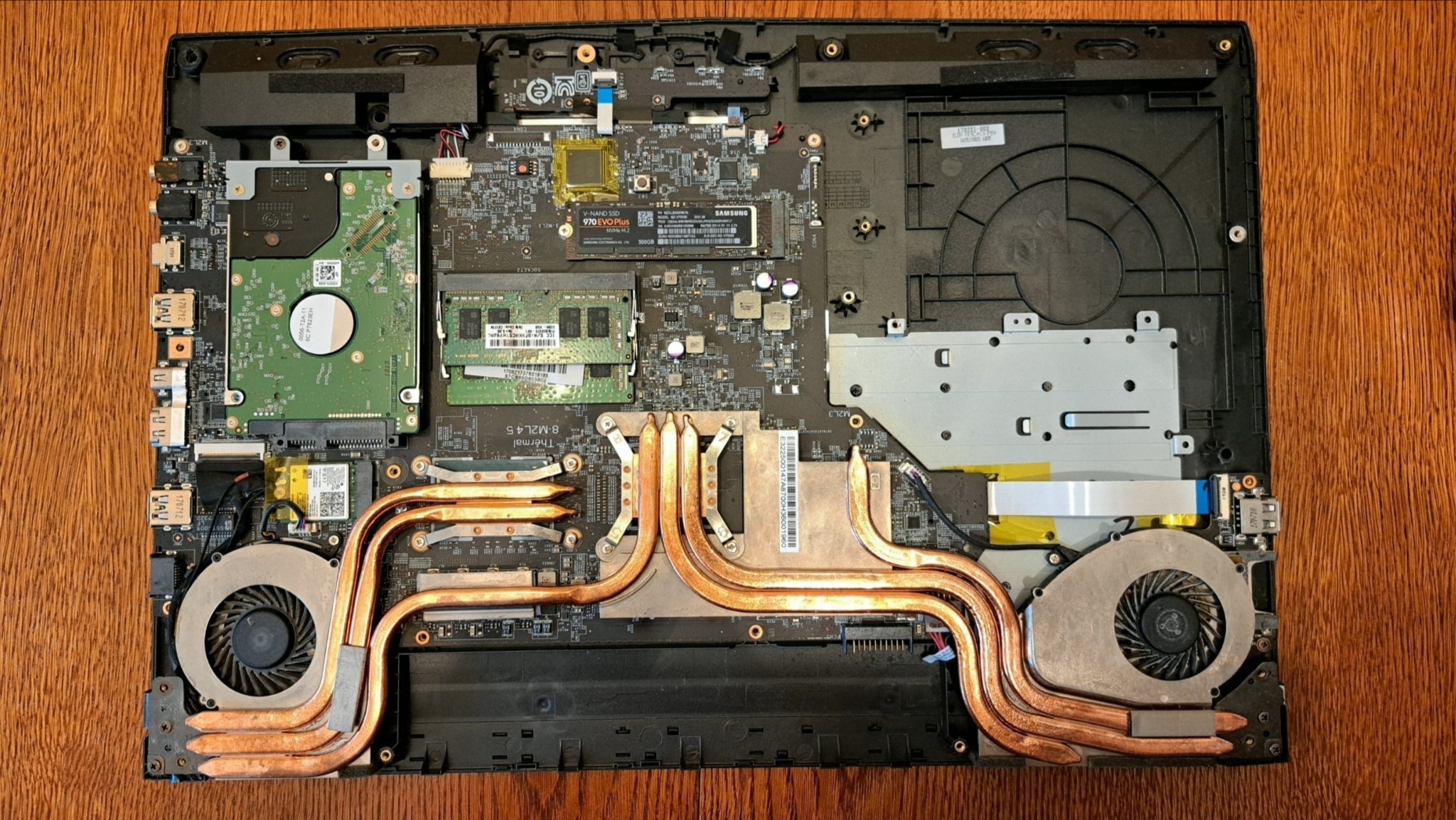 Otwarty laptop z widocznymi komponentami: dysk twardy, pamięć RAM, procesor, system chłodzenia z miedzianymi rurkami cieplnymi i wentylatory. Widok z góry na drewnianym stole.