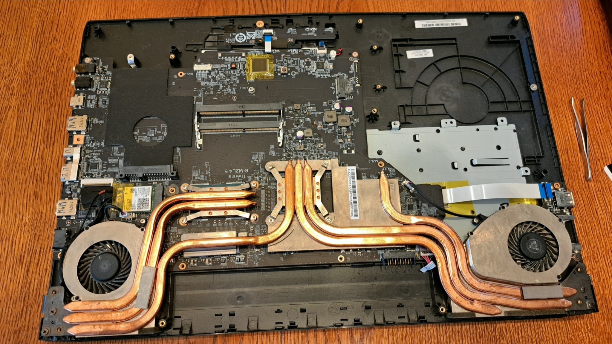 Otwarty laptop z widoczną płytą główną, systemem chłodzenia z miedzianymi rurkami cieplnymi i wentylatorami. Widoczne elementy elektroniki i narzędzia.