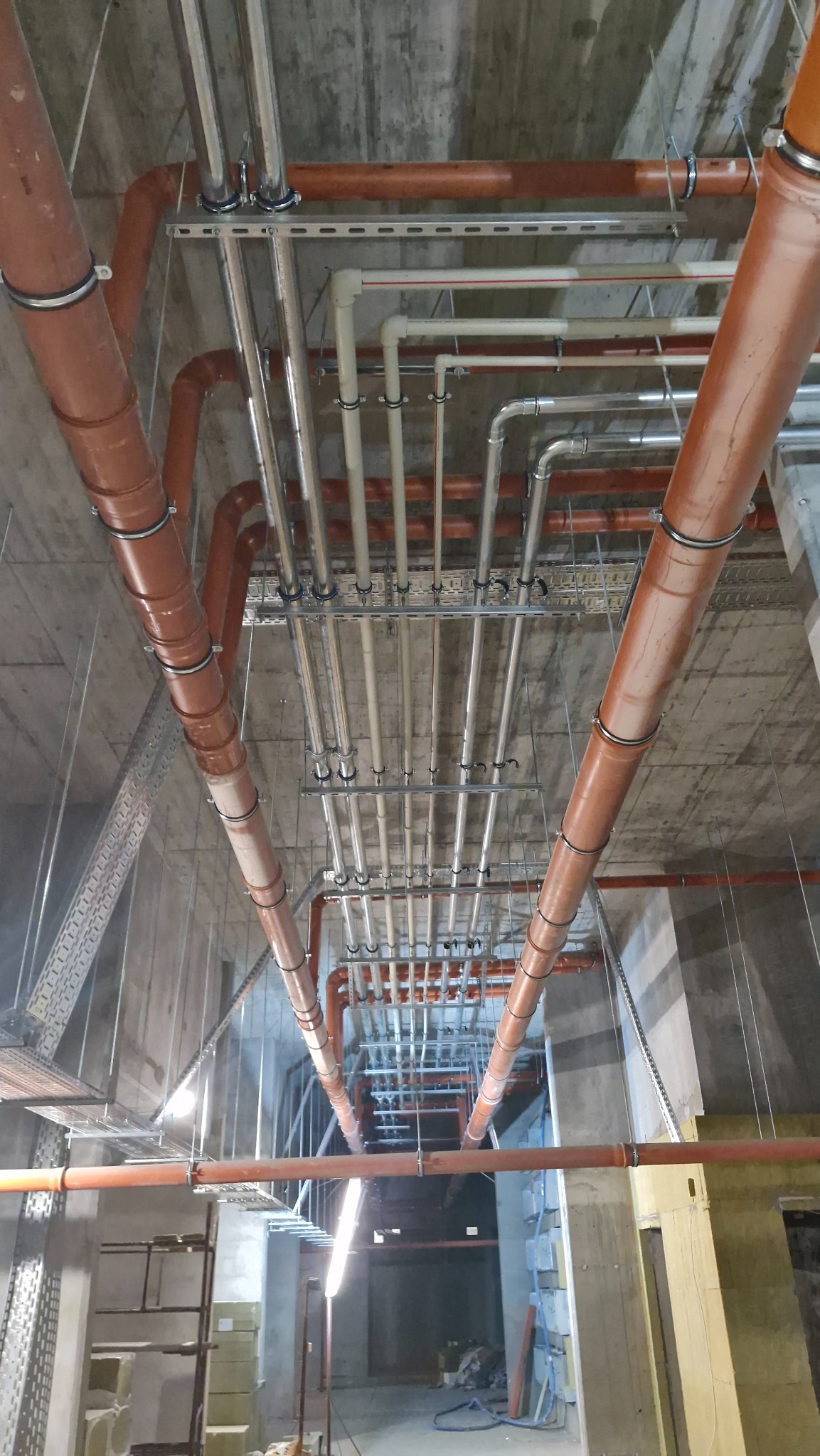 Różnokolorowe rury instalacyjne (brązowe, białe, srebrne) zamocowane pod betonowym sufitem. Widok z dołu, perspektywa zbieżna, surowy stan wykończenia.