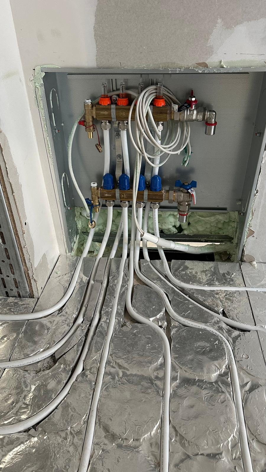 Rozdzielacz ogrzewania podłogowego z białymi rurami ułożonymi na folii aluminiowej, widok z góry, instalacja w trakcie montażu, pianka montażowa w tle.