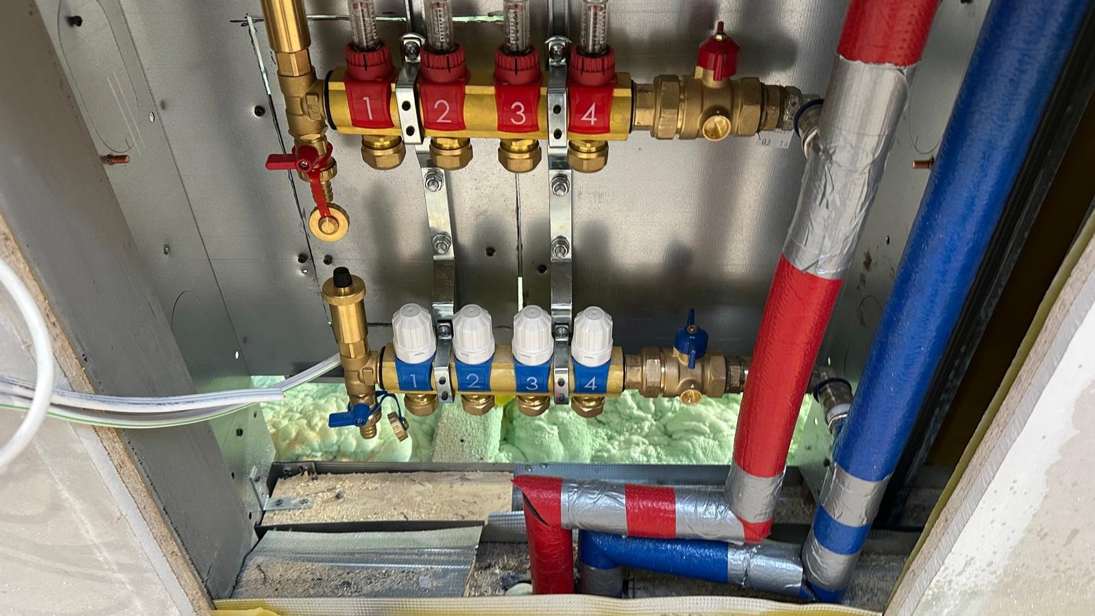 Rozdzielacz ogrzewania podłogowego z numerowanymi zaworami i rurami w izolacji, zamontowany w Toruniu. Widok z dołu na instalację.
