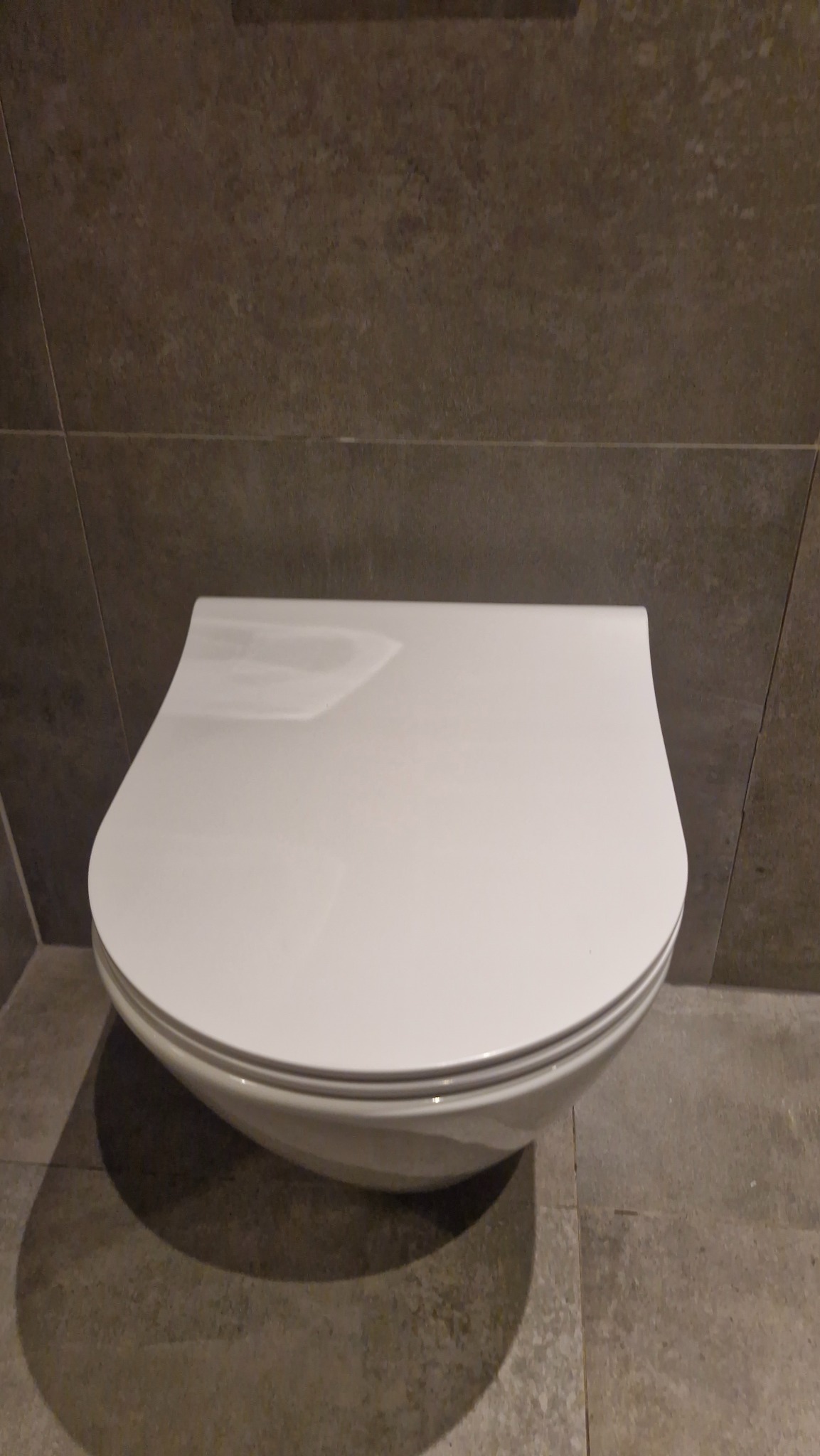 Nowoczesna, podwieszana toaleta z deską wolnoopadającą, zamontowana na tle szarych, matowych płytek. Minimalistyczny design, czysta forma.