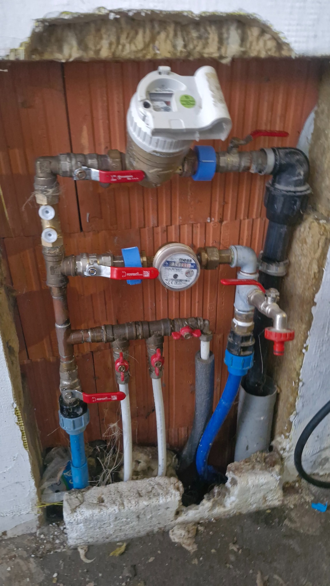 Złożona instalacja wodna z licznymi zaworami, rurami miedzianymi i plastikowymi, oraz wodomierzem, zamontowana na tle ceglanej ściany z widoczną izolacją.