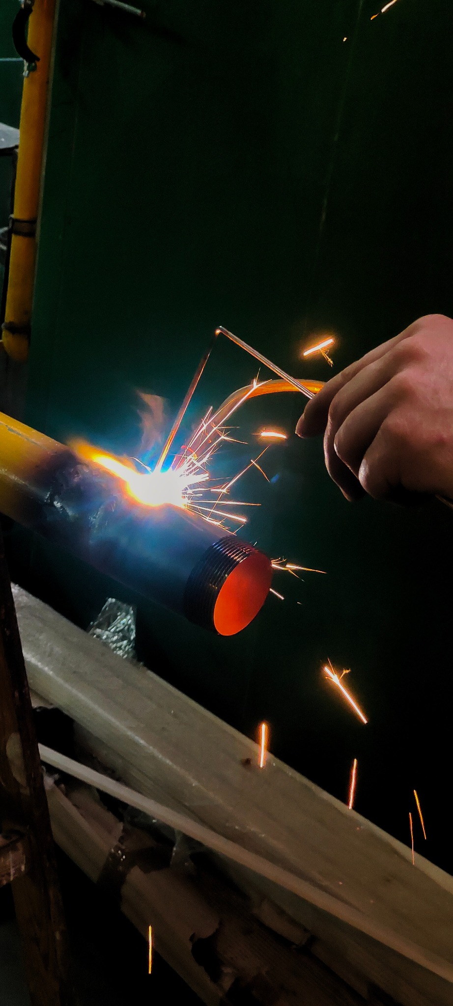 Spawanie rury z gwintem; widoczne iskry i dym. Ręka spawacza trzyma elektrodę. Żółta rura w tle. Ciemne tło. Prace spawalnicze na instalacji.
