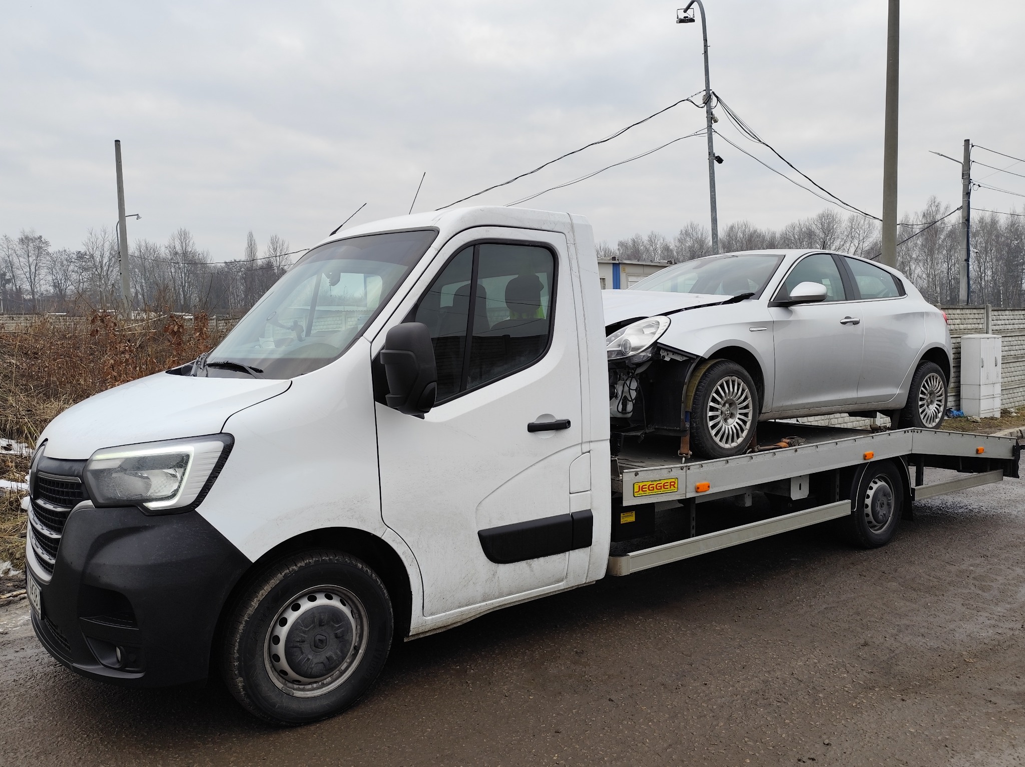 Biała autolaweta z uszkodzonym srebrnym samochodem osobowym na platformie. Widoczne uszkodzenia przodu pojazdu. Szare niebo w tle.