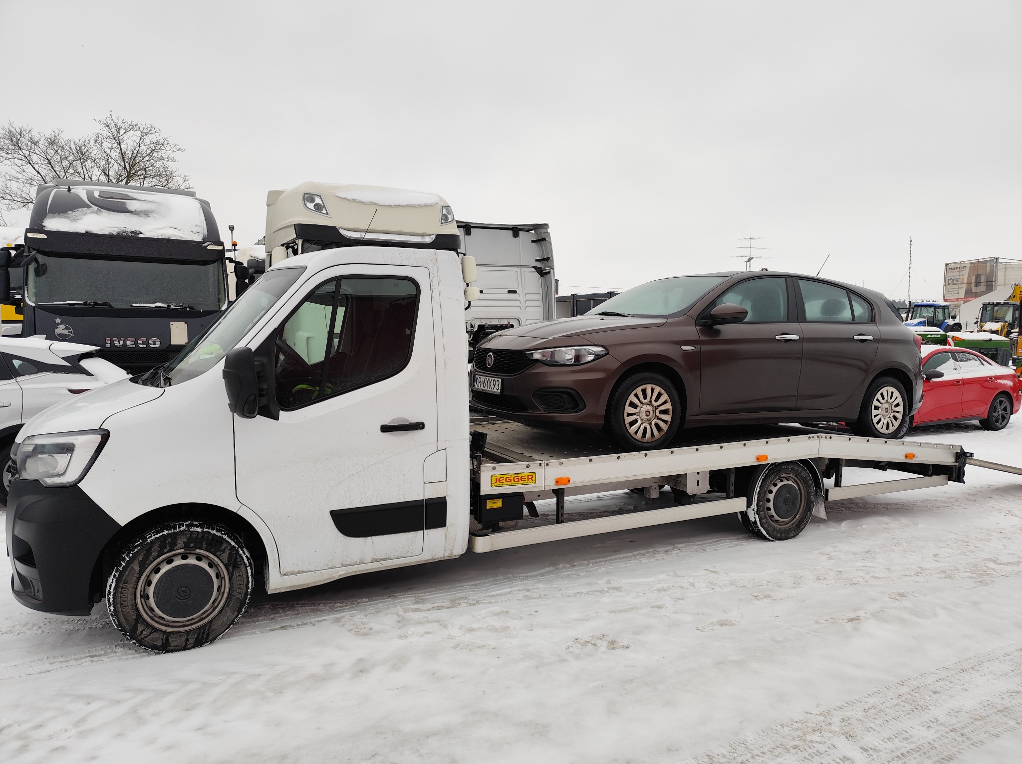 Biała autolaweta z brązowym Fiatem Tipo na platformie, zimowy krajobraz z ośnieżonymi pojazdami w tle. Widoczne detale: opony zimowe, tablica rejestracyjna.