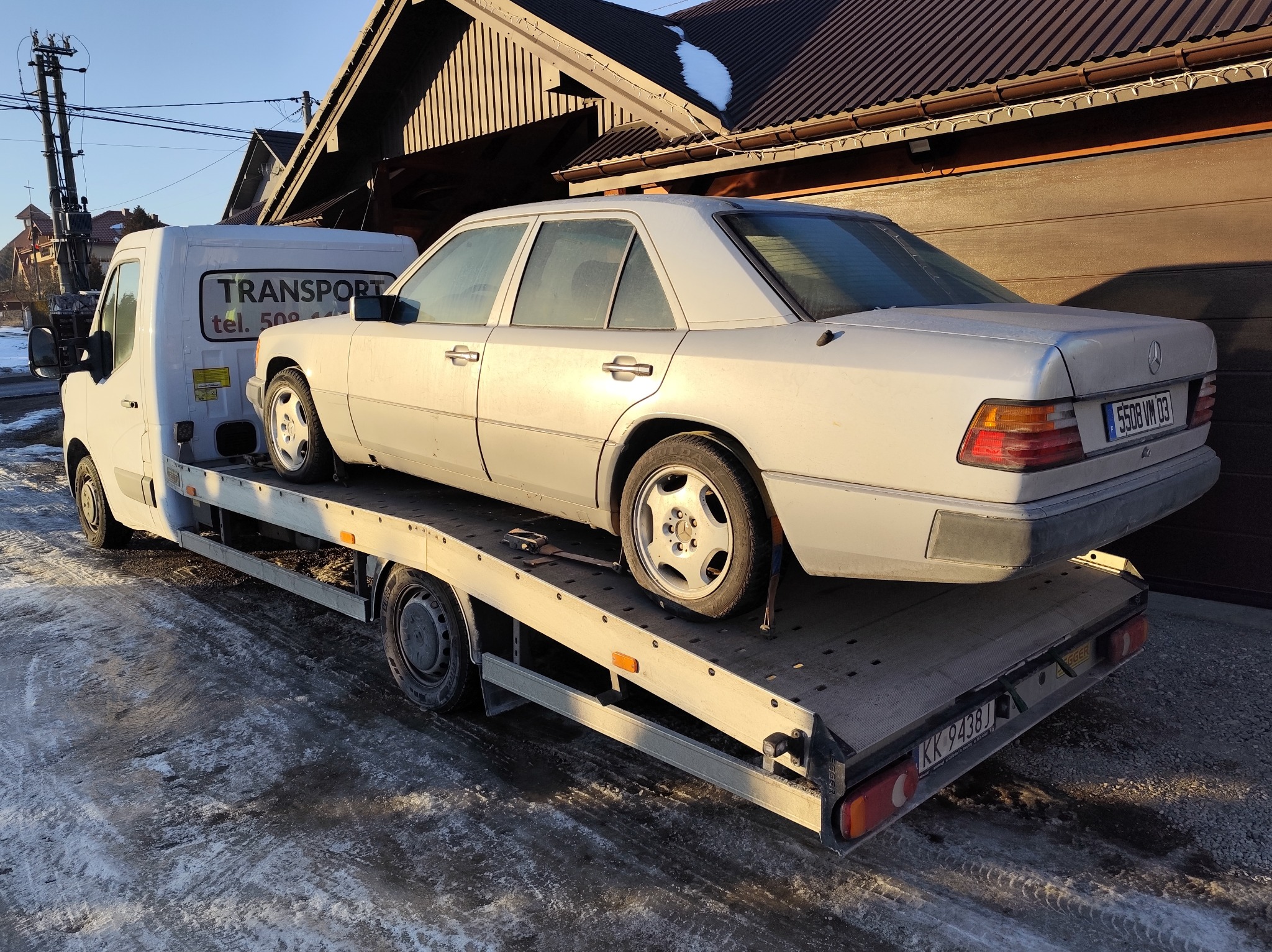 Biała laweta z zaparkowanym na niej białym Mercedesem W124 na tle budynku z brązowym dachem. Widoczne tablice rejestracyjne pojazdów i logo firmy transportowej.