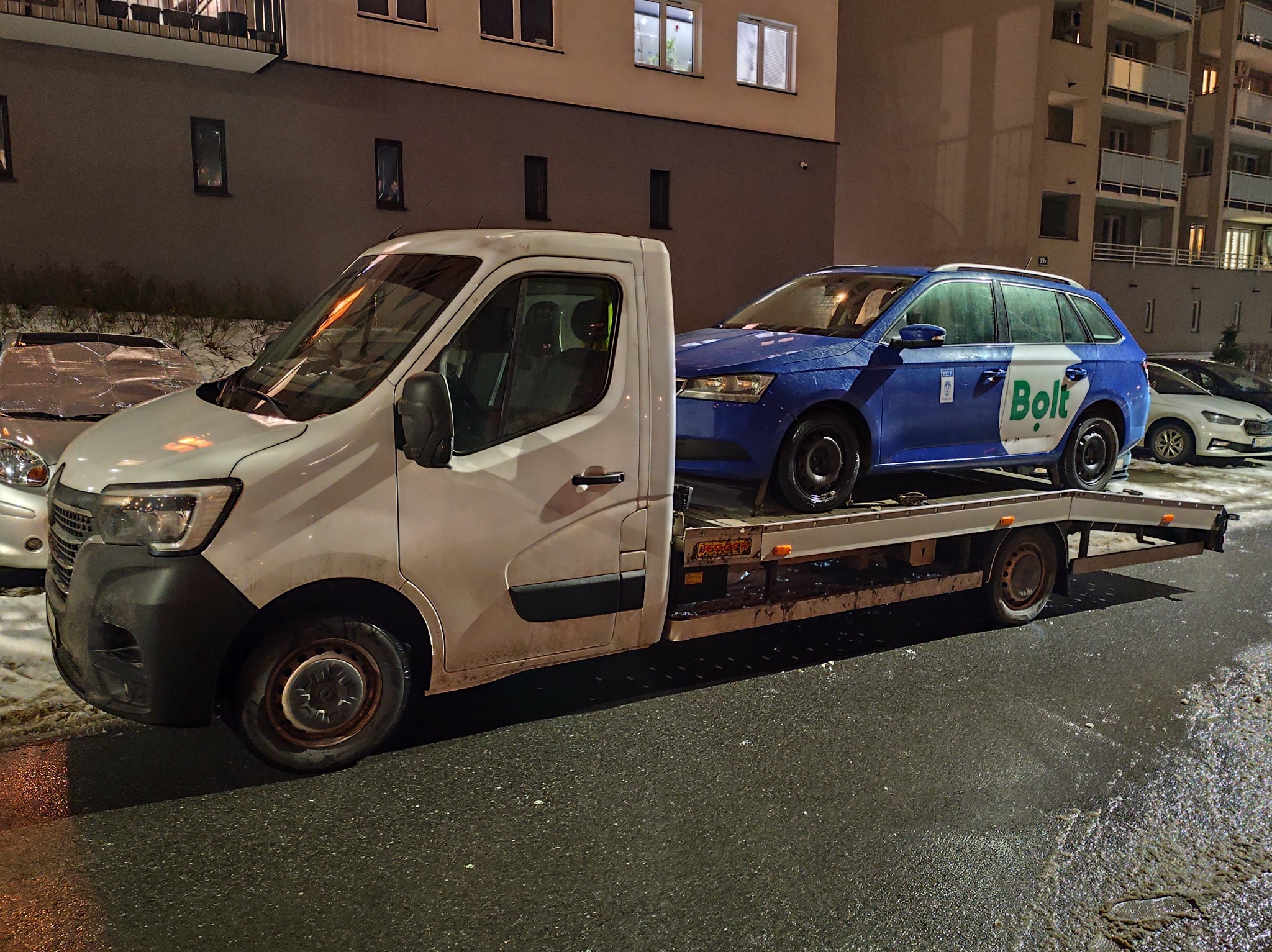 Biała platforma transportowa z niebieskim samochodem Bolt na lawecie, zaparkowana na mokrej ulicy w nocy, z budynkami mieszkalnymi w tle.