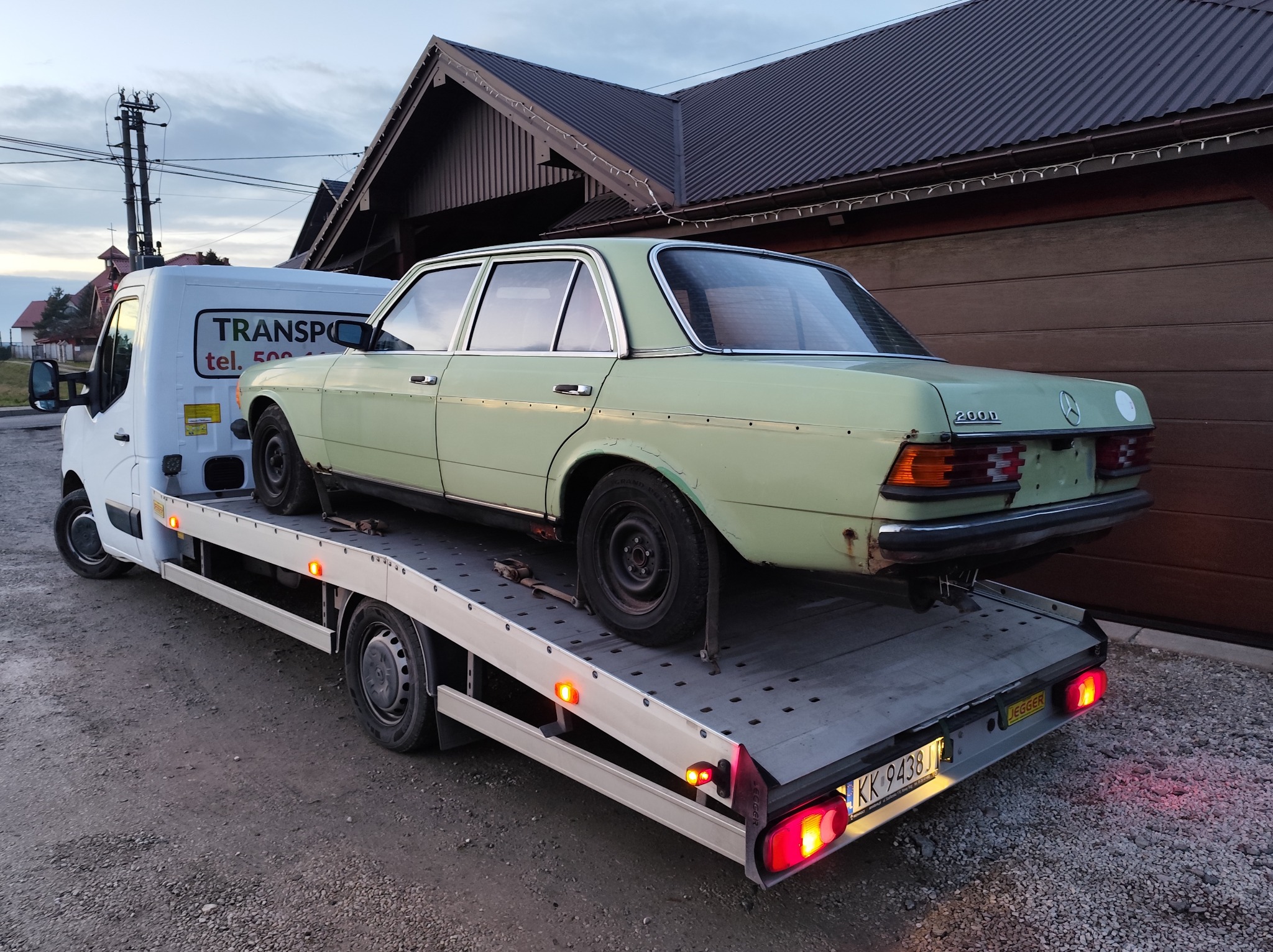 Laweta z załadowanym, jasnym Mercedesem 200D na tle budynku z ciemnym dachem. Widoczny fragment białego busa z napisem 'TRANSPORT'.