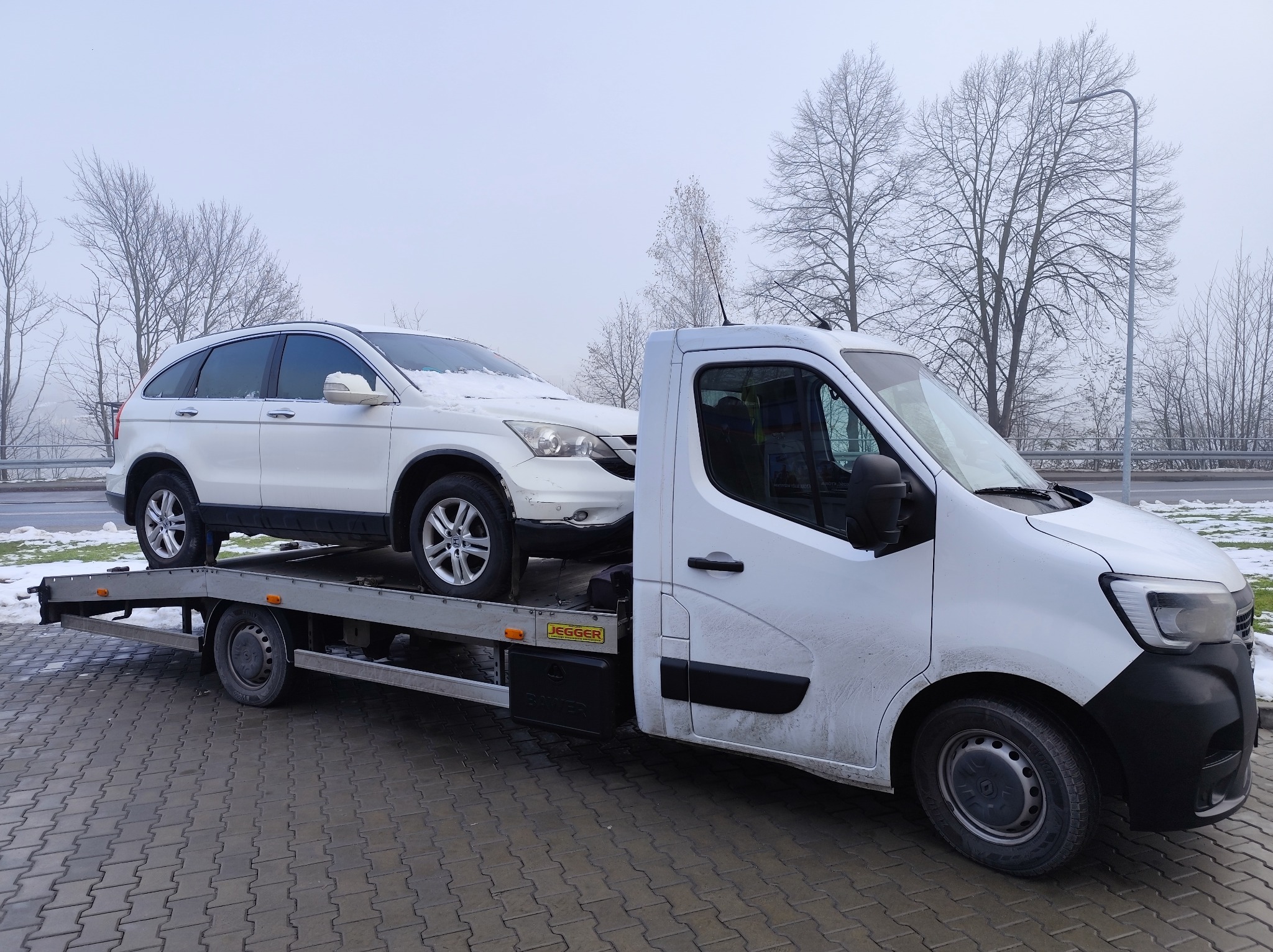 Biały samochód osobowy, częściowo pokryty śniegiem, transportowany na platformie białej ciężarówki w pochmurny dzień. Widoczne drzewa w tle.
