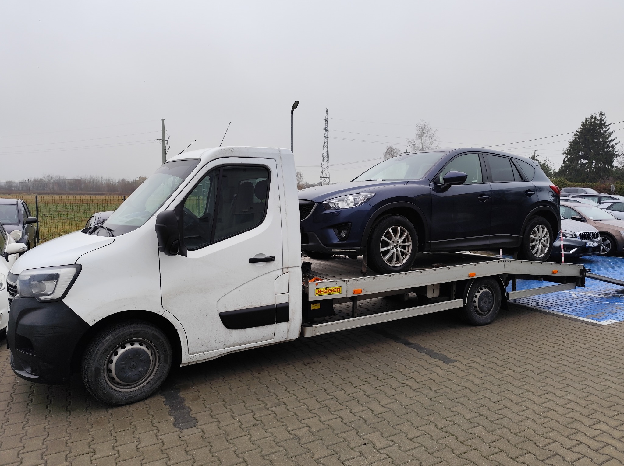 Biała ciężarówka z platformą transportową przewozi granatowy samochód osobowy na parkingu. Widoczne inne pojazdy i słup wysokiego napięcia w tle.