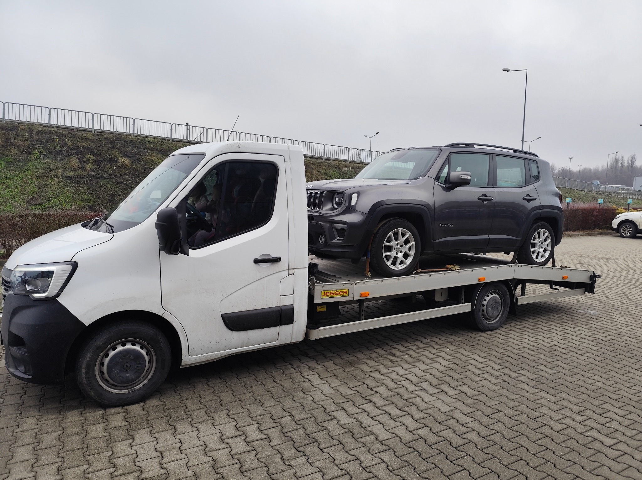 Biała laweta z szarym Jeepem Renegade na platformie, zaparkowana na brukowanym placu w pochmurny dzień. Widoczny fragment drogi i barierki w tle.